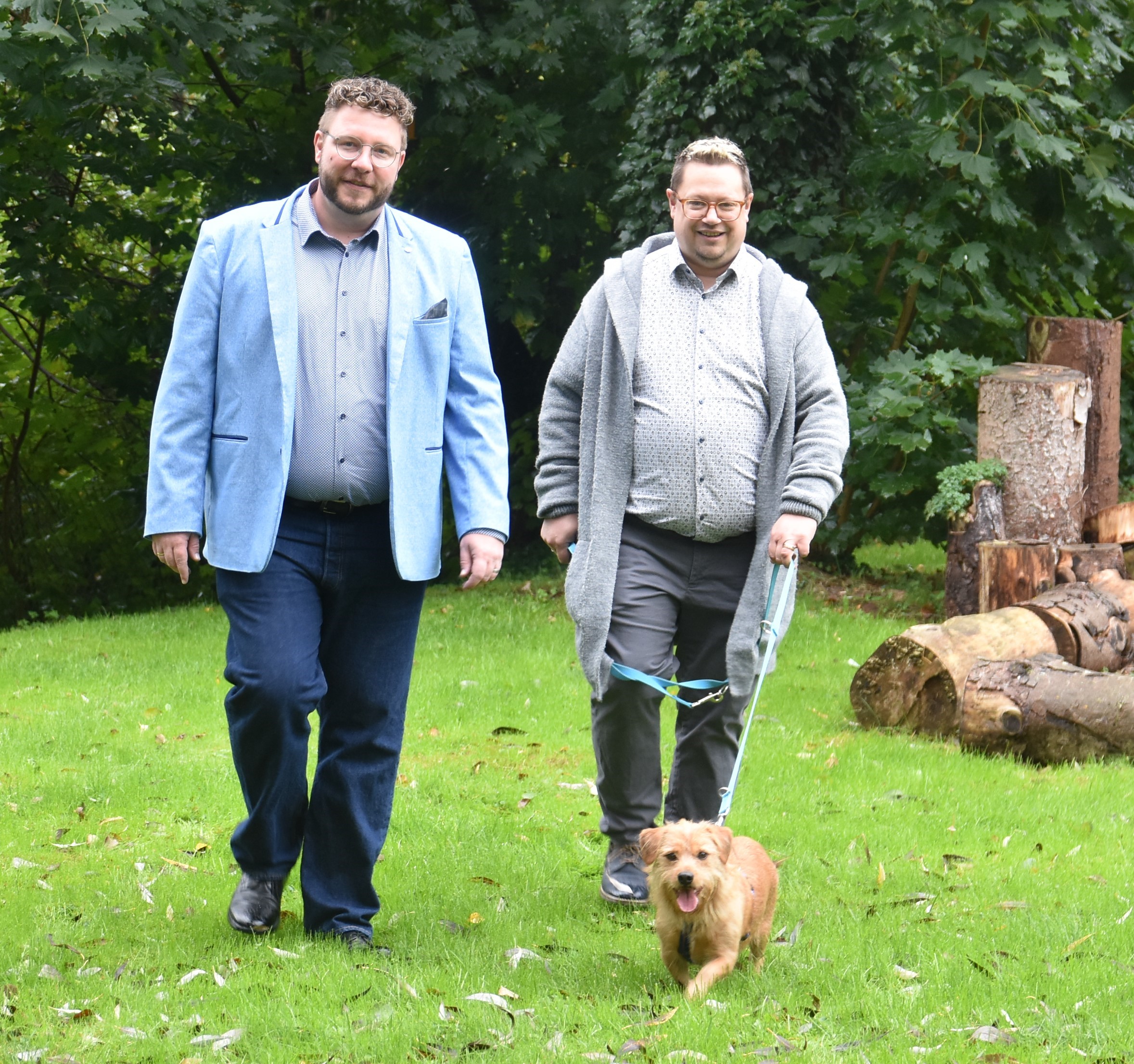 Vikar Tobias Vonderlehr und sein Ehemann Nik laufen gemeinsam mit ihrem kleinen Hund Romeo durch einen Garten.