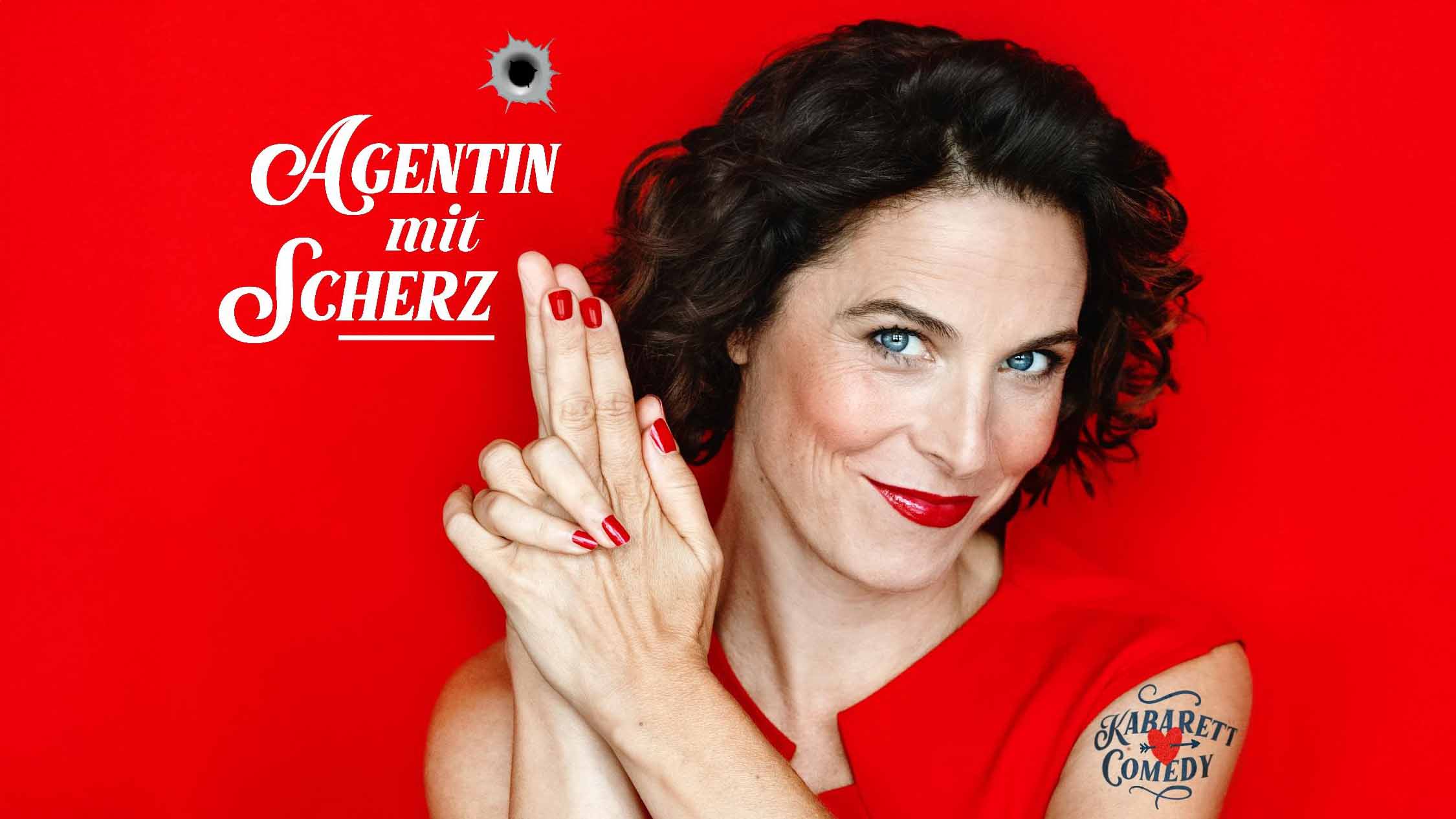 Katalyn_Hühnerfeld_C_Jeanne_Degraa_16zu9.jpg Frau in Rot mit dunklen Locken hält ihre Hände wie eine Pistole; daneben steht „Agentin mit Scherz“, darüber ein Einschussloch.
