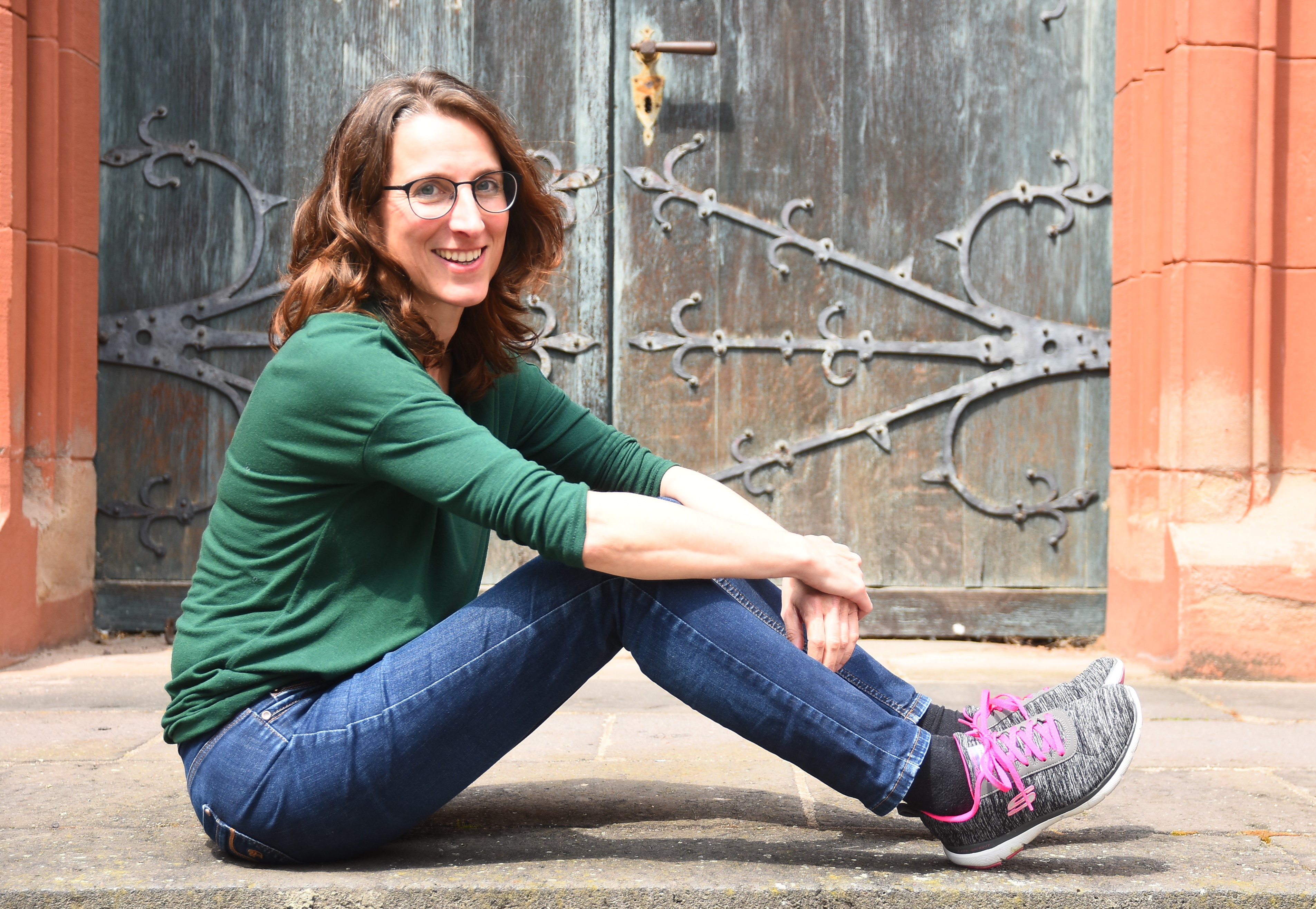 Antje Armstroff, in Jeans und T-Shirt, sitzt auf den Treppenstufen vor einem Kirchenportal. Ihre Arme umfassen ihre Beine.