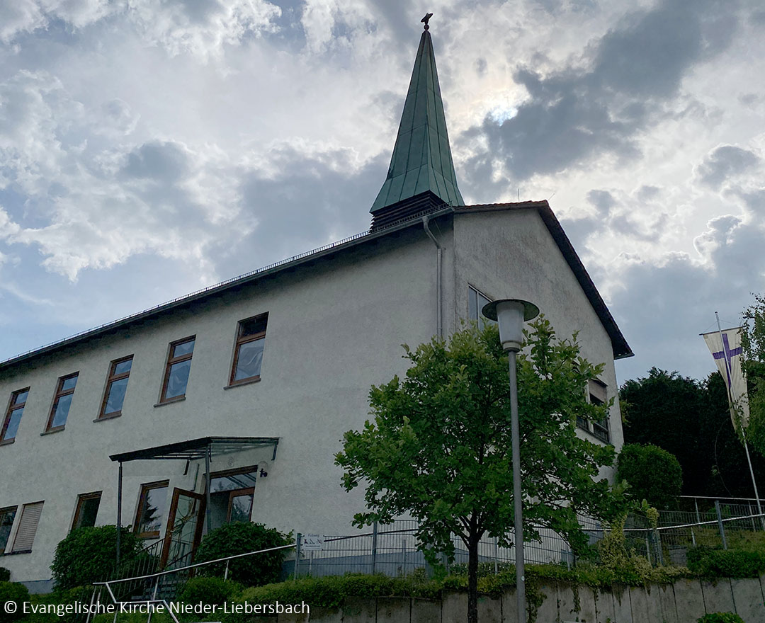 Außenansicht der Evangelischen Kirche Nieder-Liebersbach. Das schlichte, weiß verputzte Gebäude besitzt ein langgezogenes Satteldach und einen schlanken, grün gedeckten Kirchturm mit Kreuz. Vor dem Eingang stehen ein Laubbaum und eine Straßenlaterne. Im Hintergrund ziehen dichte Wolken über den Himmel.