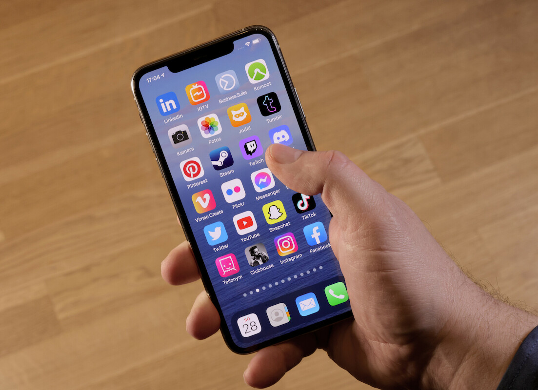 Man sieht ein Handy/IPhone in einer Hand mit den verschiedensten Apps für Socialmedia vor einem Holzhintergrund.