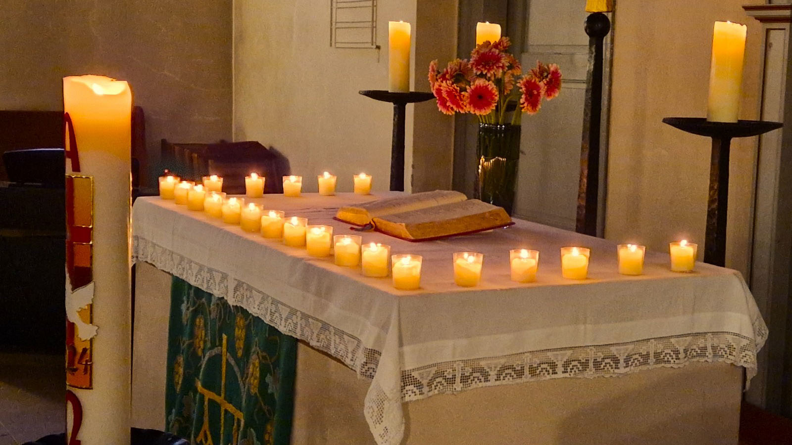 Kerzen beleuchten einen Altar mit aufgeschlagener Bibel; im Hintergrund stehen Blumen und weitere Kerzen in einem ruhigen Kirchenraum.