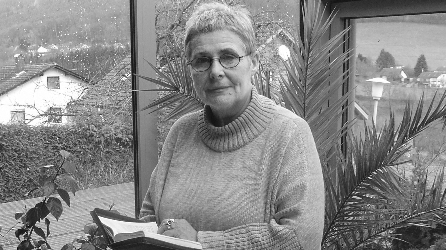 Ältere Frau mit Kurzhaarschnitt und Brille steht in einem Wintergarten und hält ein aufgeschlagenes Buch in den Händen, Pflanzen und Häuser im Hintergrund.