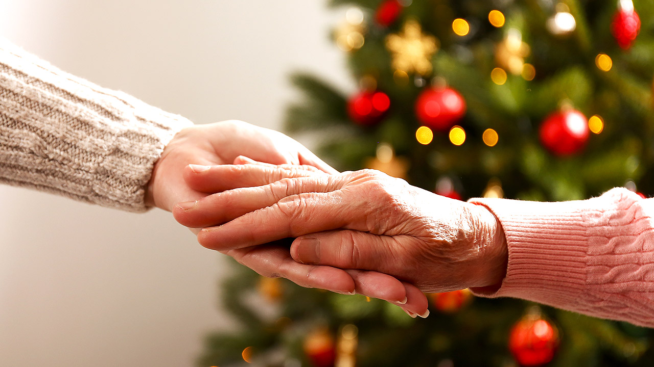 Eine Hand hält die Hand einer älteren Dame, dahinter ein geschmückter Weihnachtsbaum