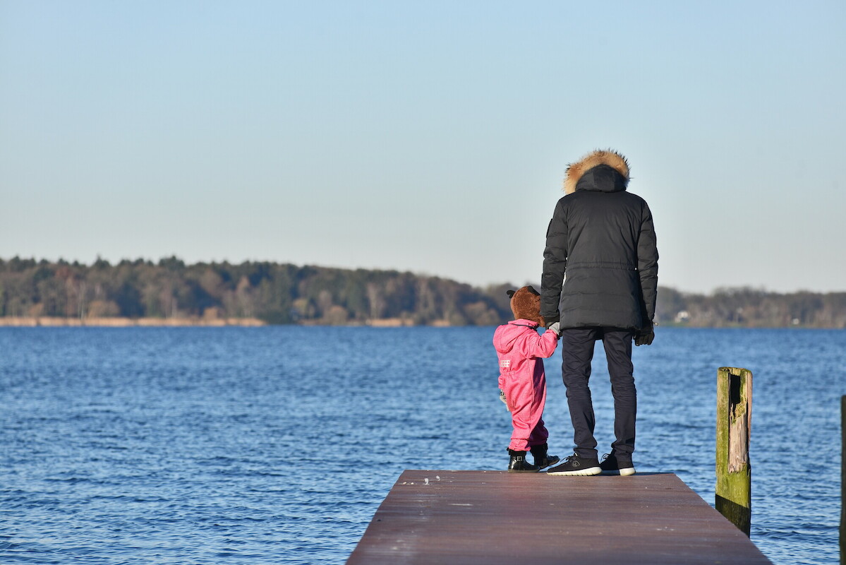 Ein Vater mit seinem Kind Hand in Hand auf einem Steg über Wasser.
