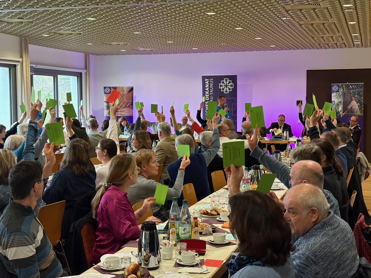 Raum mit vielen Tischen und Stühlen, Menschen heben die Hände mit einer grünen Karte in der hand und stimmen ab.