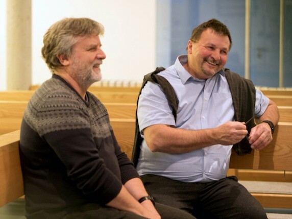 Zwei Männer im ALter von etwa 45 bis 55 Jahre sitzen entspannt in legerer Kleidung auf Kirchenbänken und lächeln