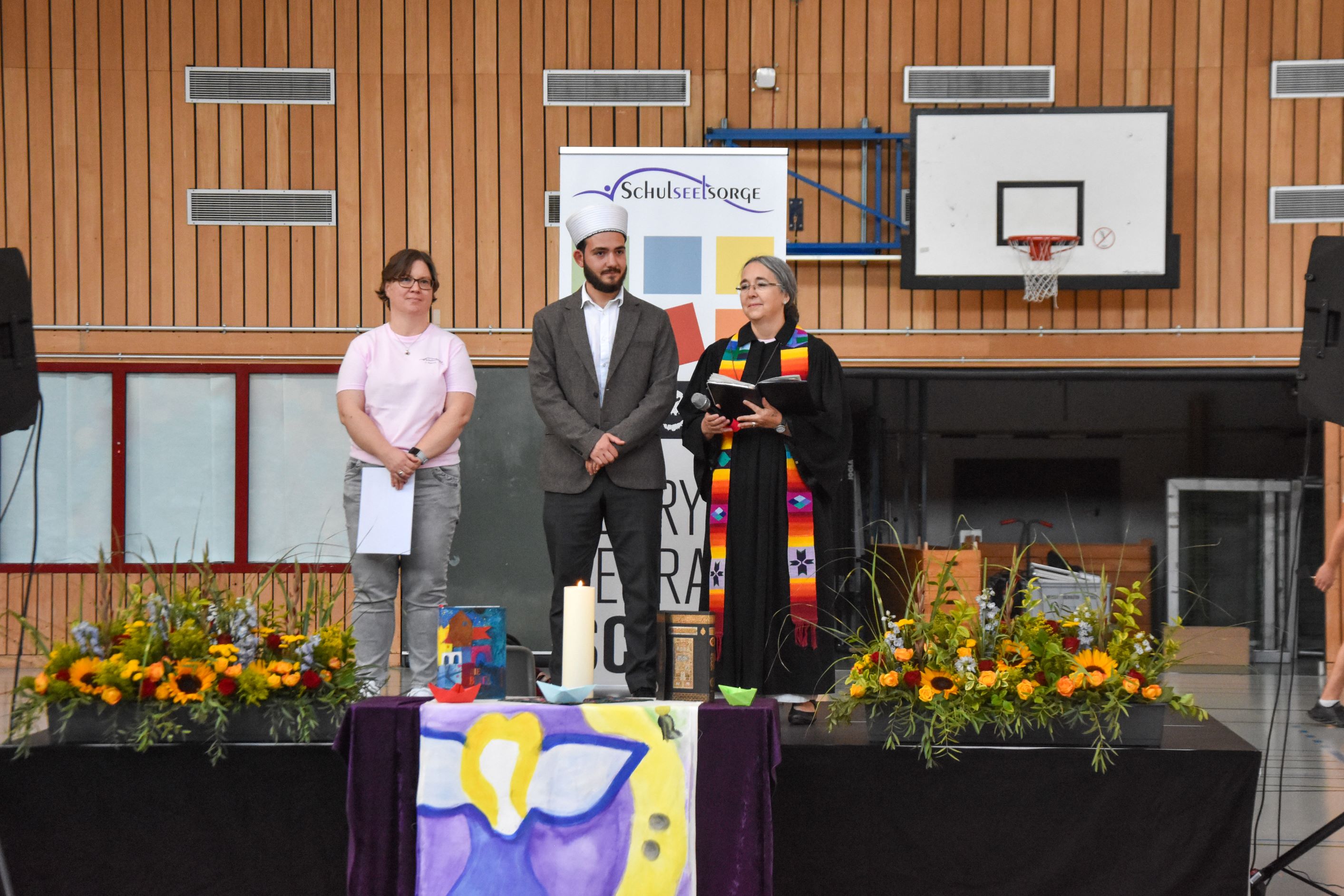 Drei Personen auf einer Bühne in einer Turnhalle.