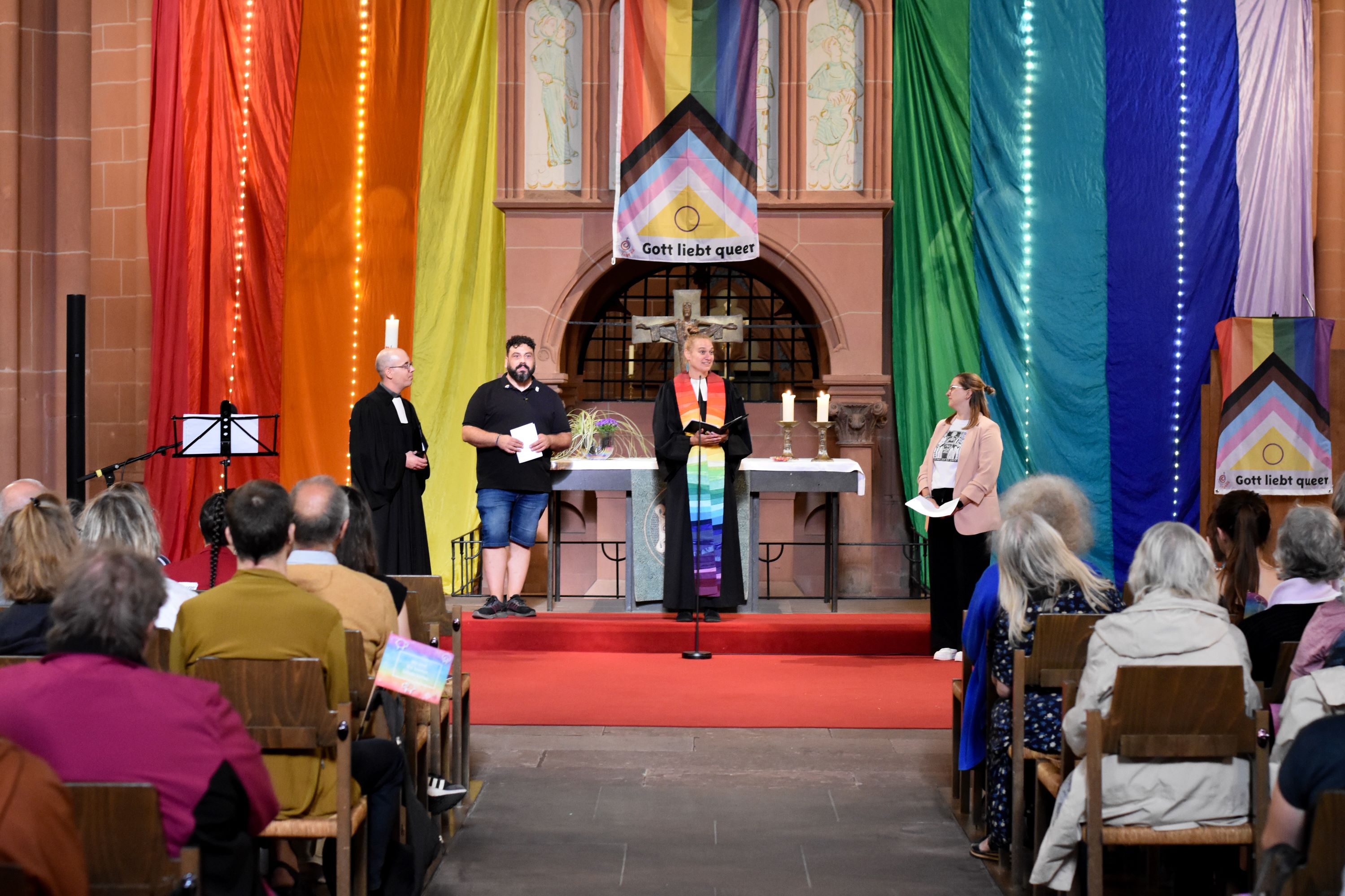 Der mit Regenbogenflagge geschmückte Altarraum einer Kirche. Vor dem Altar stehen vier Menschen