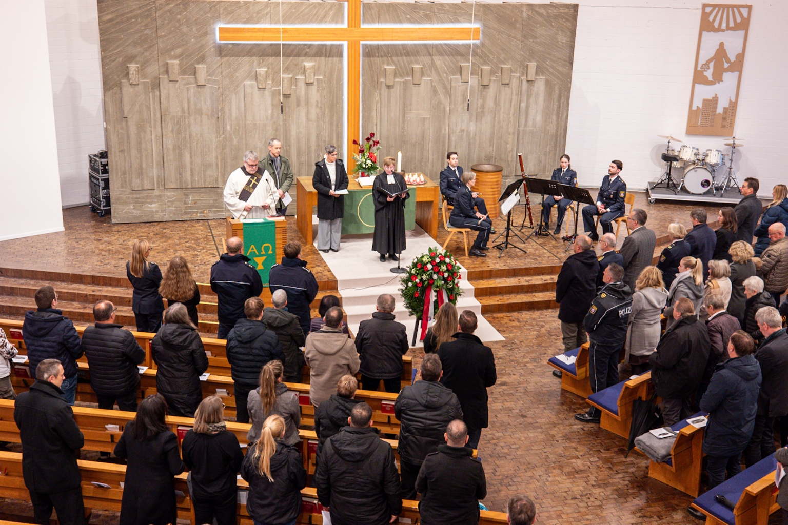 Im Inneren der Paul-Gerhardt-Kirche stehen die Besucher in den Reihen. Im Altarraum stehen die Pfarrerin, der Diakon und weitere Beteiligte. Das kleine Orchester sitzt rechts.