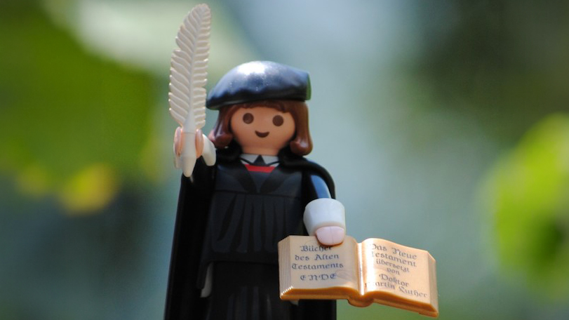 Playmobil-Figur von Martin Luther in schwarzer Gelehrtenkleidung mit Federkiel und aufgeschlagenem Buch in den Händen.