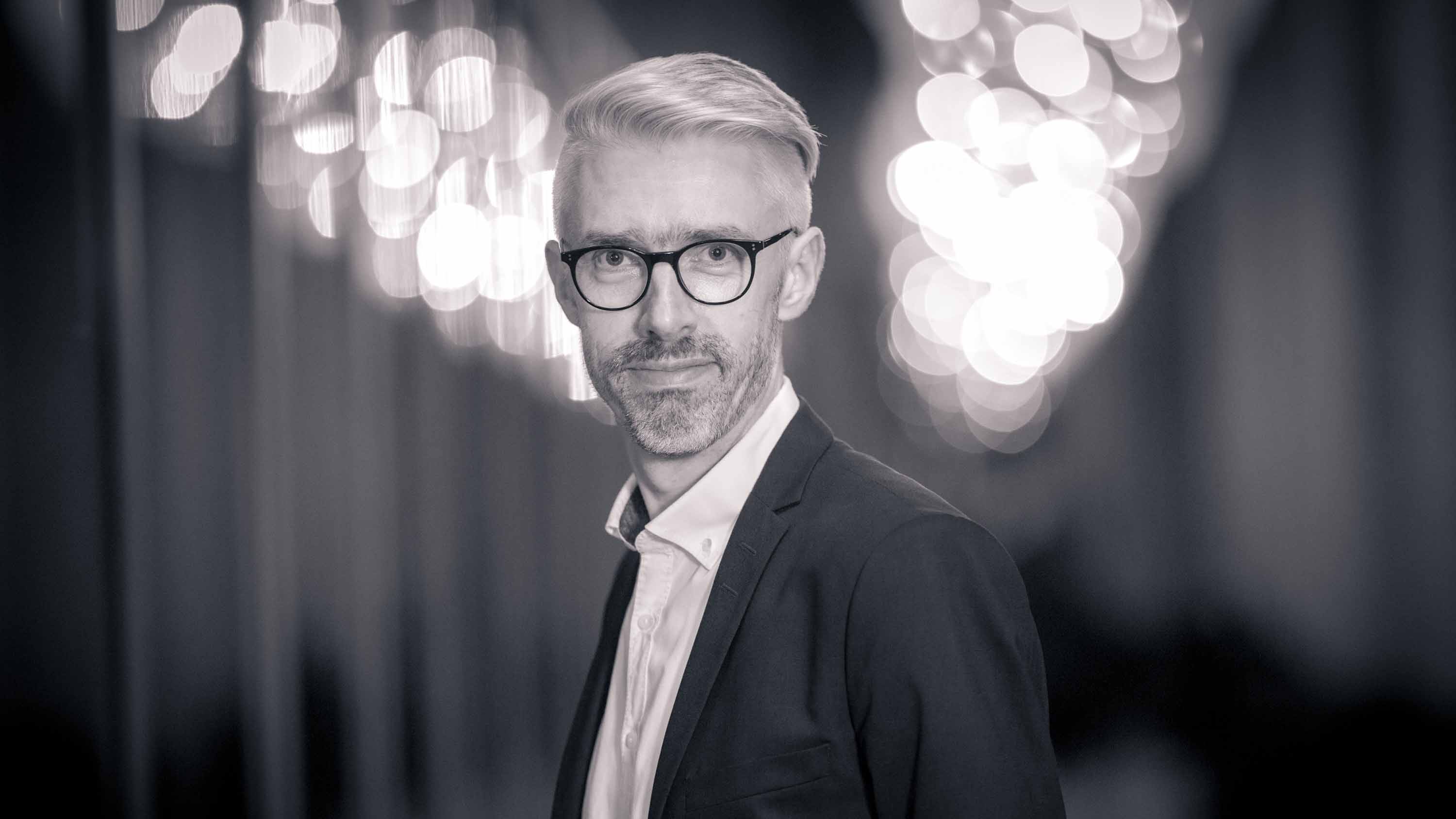 Porträt einer Person mit heller Kurzhaarfrisur und Brille im dunklen Sakko und weißen Hemd, die frontal in die Kamera blickt; unscharfe Lichter im Hintergrund.