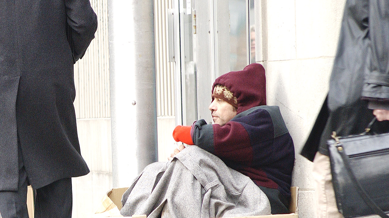 Obdachloser Mann sitzt im Winter in einer Fußgängerzone, Menschen eilen vorbei