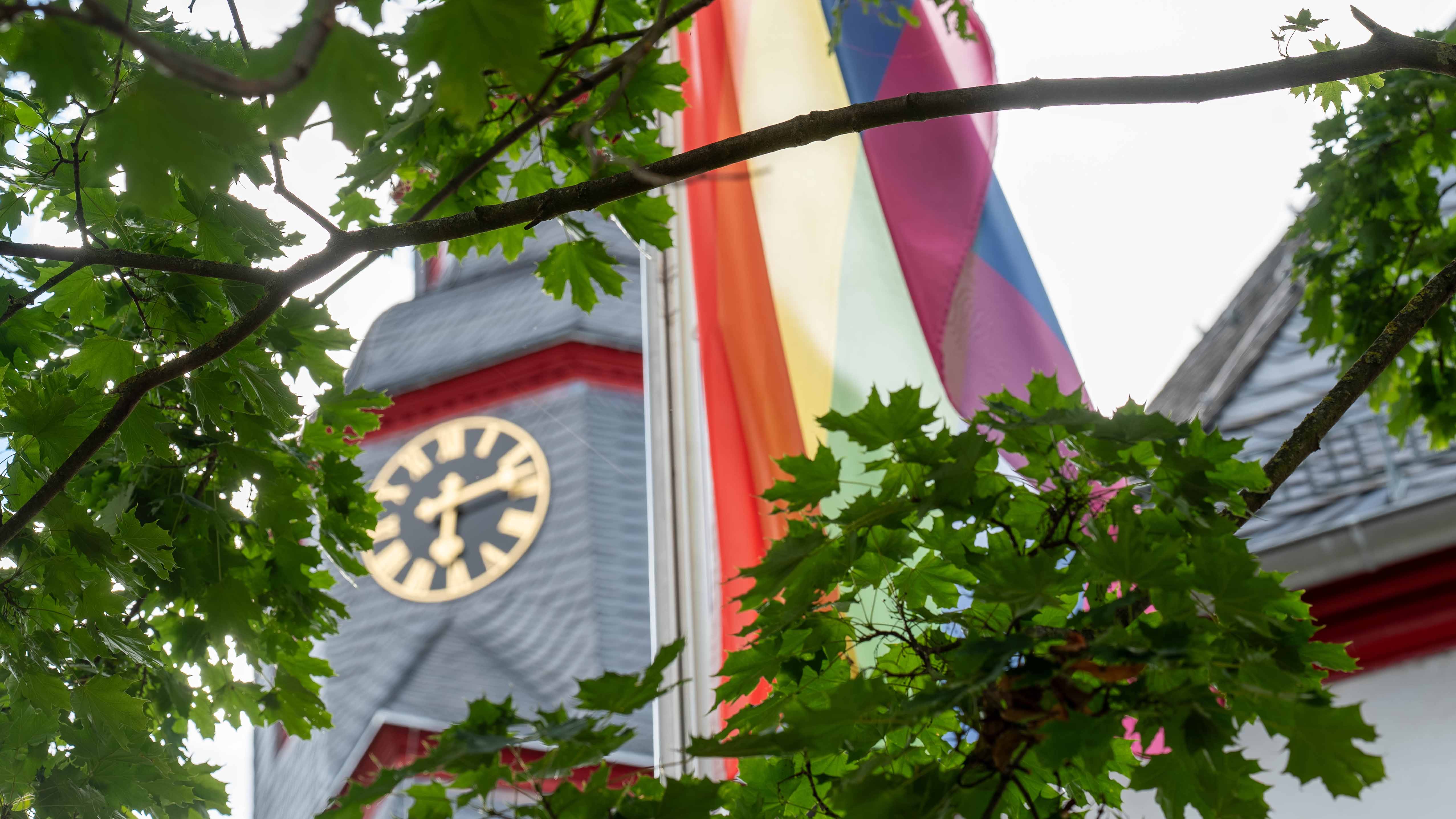 Kirchturm mit Uhr und einer gehissten Pride-Flagge, eingerahmt von grünen Baumzweigen vor hellem Himmel.
