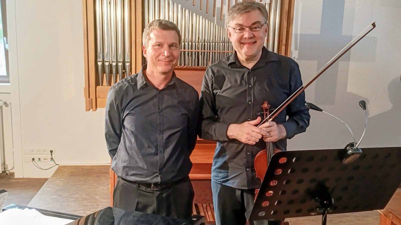Rolf Fritz (links) und Alexander Galushkin (rechts) stehen lächelnd vor einer Kirchenorgel. Beide tragen dunkle Hemden. Galushkin hält eine Violine und einen Geigenbogen in den Händen. Im Vordergrund ist ein schwarzer Notenständer zu sehen.