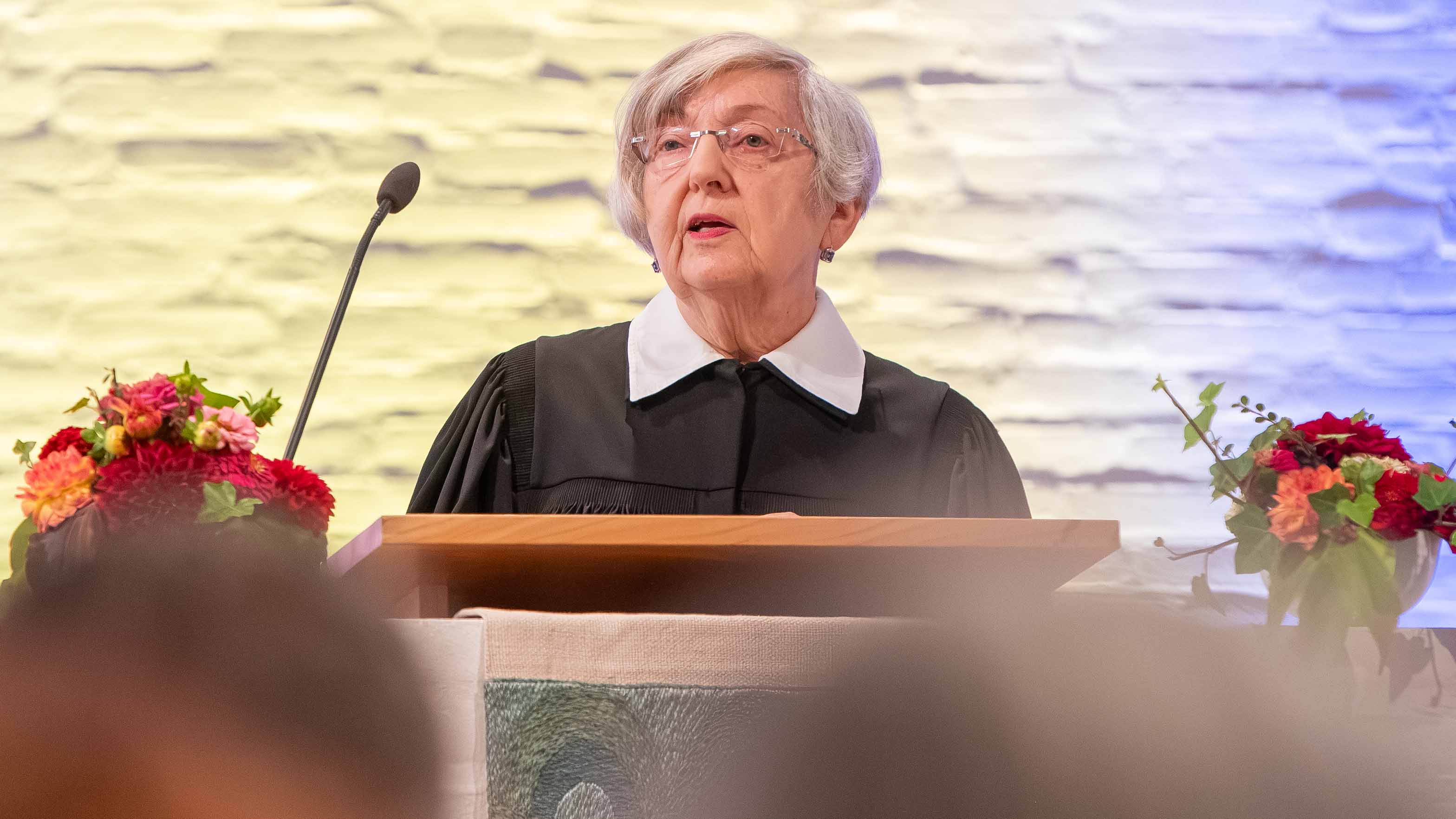 Eine ältere Frau mit kurzen grauen Haaren und Brille steht im schwarzen Talar mit weißem Kragen hinter einem hölzernen Lesepult und spricht in ein Mikrofon. Links und rechts rahmen farbenfrohe Blumenarrangements die Szene ein. Im Hintergrund ist eine hell beleuchtete Natursteinwand zu sehen.