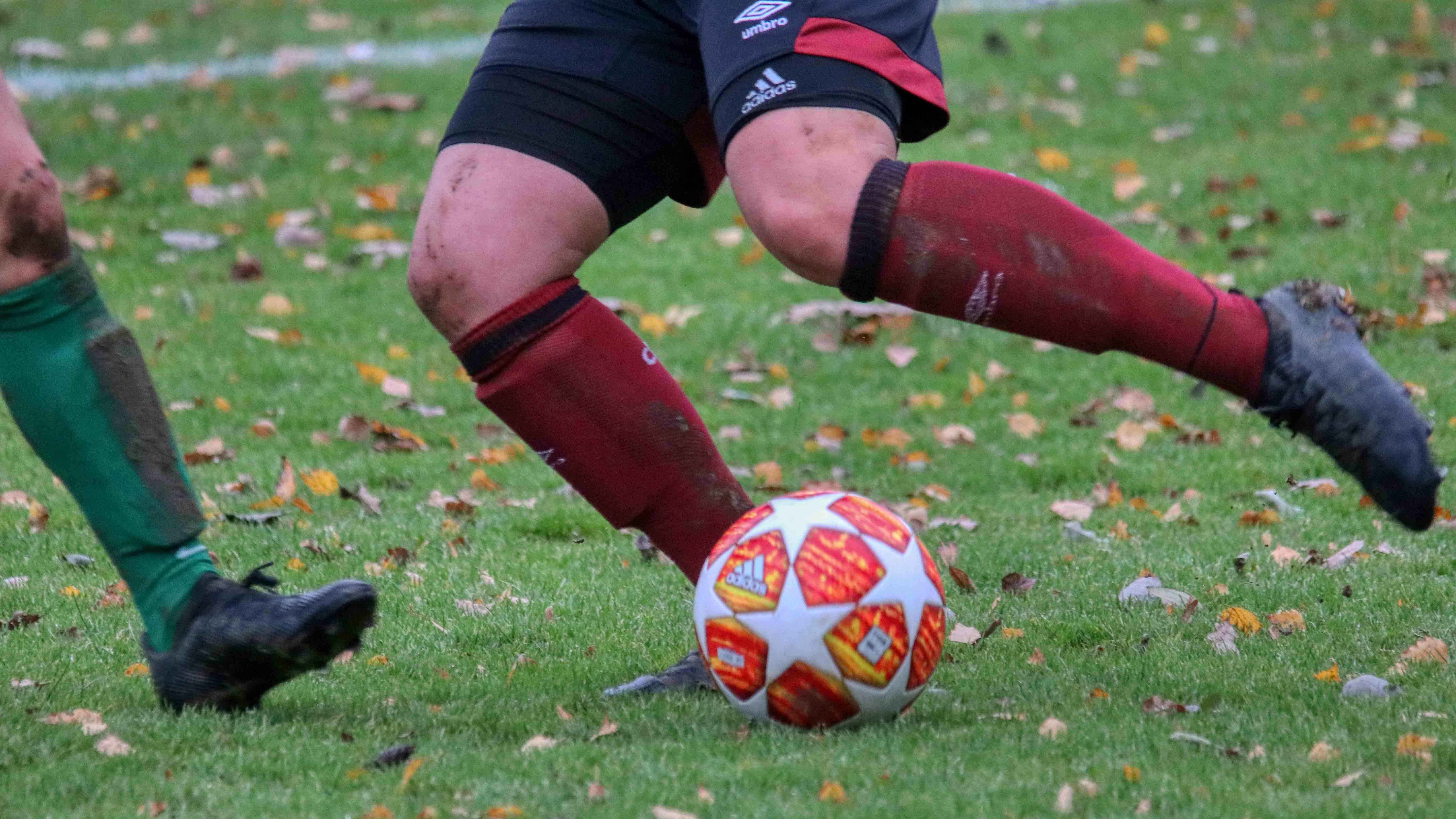 Zwei Fußballspieler kämpfen auf einem Rasen um den Ball; sichtbar sind nur ihre Beine, ein Spieler führt den Ball, der andere greift von links an.