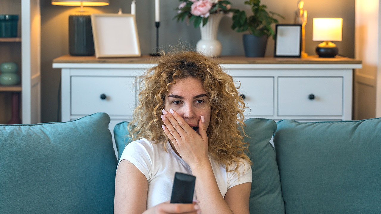 Eine Frau auf der Couch mit Fernbedienung ist erschrocken über die aktuelle Nachrichtenlage