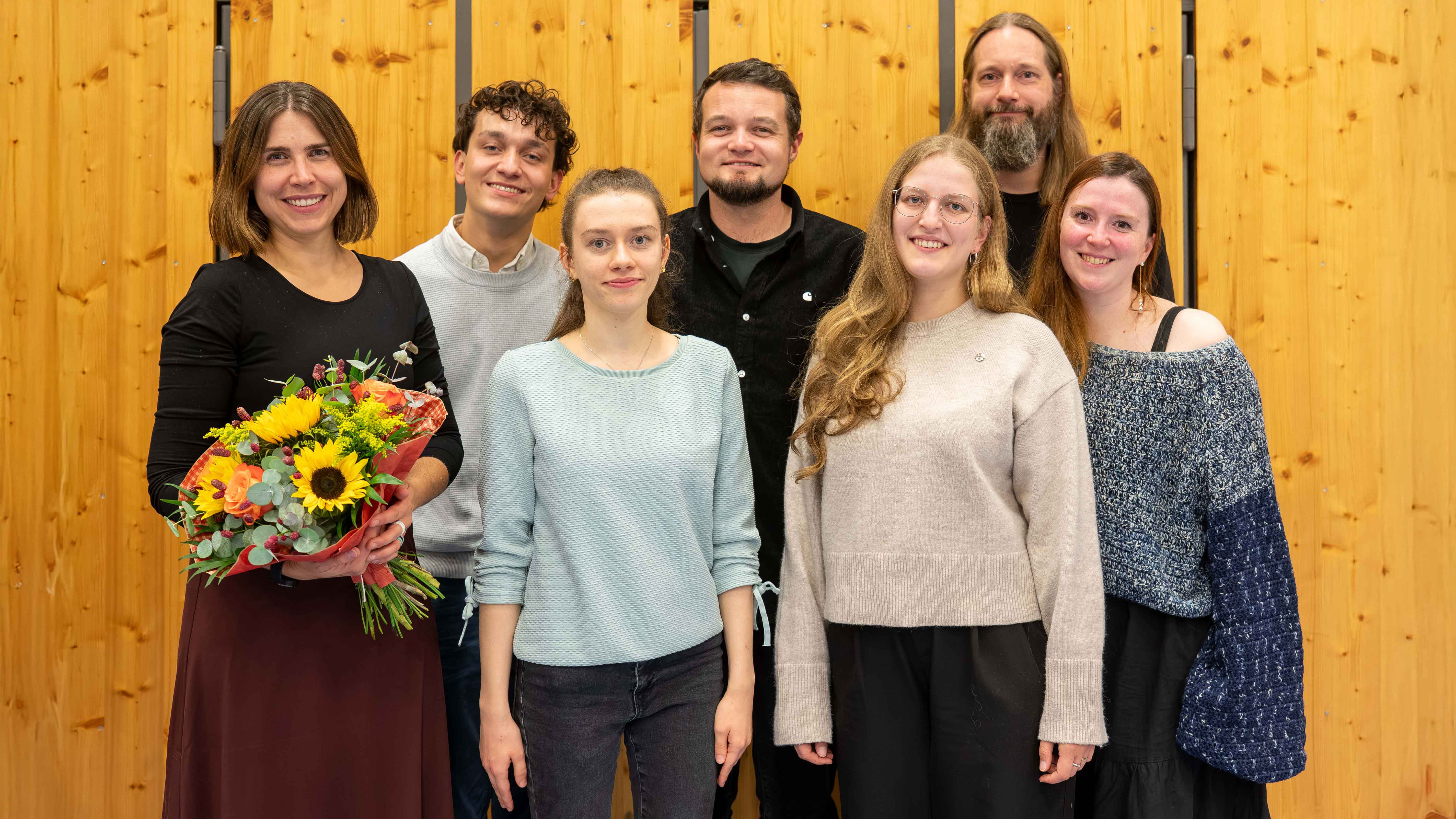 Gruppenfoto von sieben lächelnden Personen vor einer hellen Holzwand. Links steht eine Frau in schwarzem Oberteil und dunkelrotem Rock, die einen großen Blumenstrauß mit Sonnenblumen hält. Neben ihr stehen drei Frauen und drei Männer in legerer Kleidung. Ein großer Mann mit langen Haaren und Vollbart steht in der hinteren Reihe.