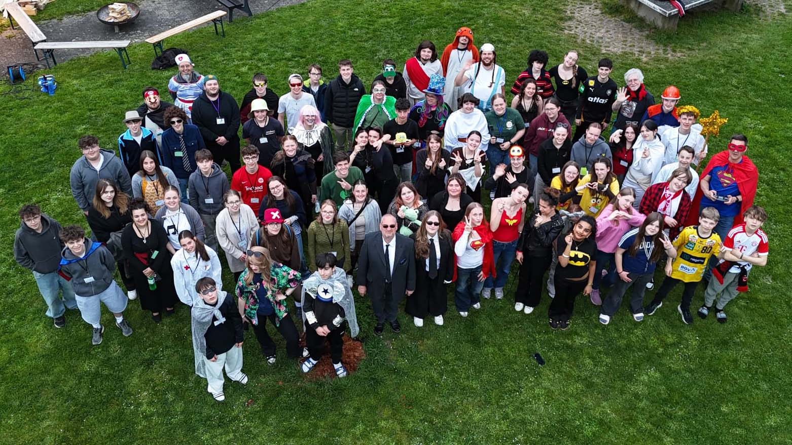 Große Gruppe junger Menschen in Kostümen und Freizeitkleidung steht auf einer Wiese und blickt für ein Gruppenfoto nach oben. 