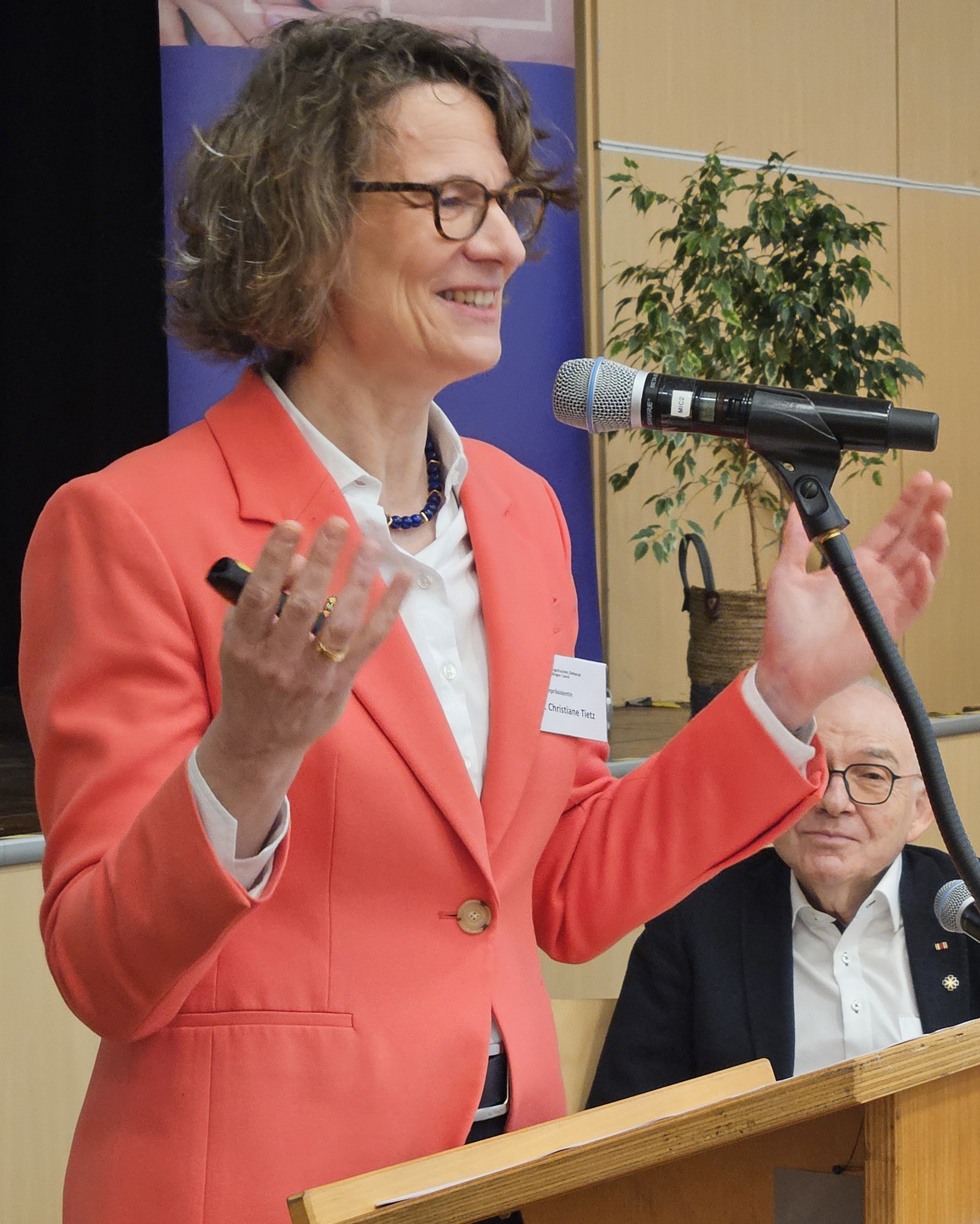 Christiane Tietz am Rednerpult mit leicht erhobenen, offenen Armen, vor sich ein Mikrofon. Sie trägt einen lachsfarbenen Blazer und lächelt. Im Hintergrund erkennt man Präses Rolf Hartmann und eine Grünpflanze auf der Bürgerhaus-Bühne.