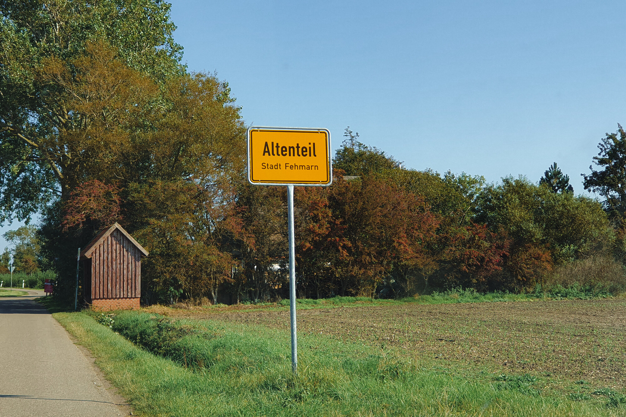 Man sieht eine Wiese eine Hecke und einen Straßenrand, über den sich die Äste eines großen Baumes neigen, im Hintergrund ein sehr kleines Holzhäuschen. In der Mitte des Bildes sieht man ein gelbes Straßenschild mit der Aufschrift "Altenteil - Stadt Fehmarn".
