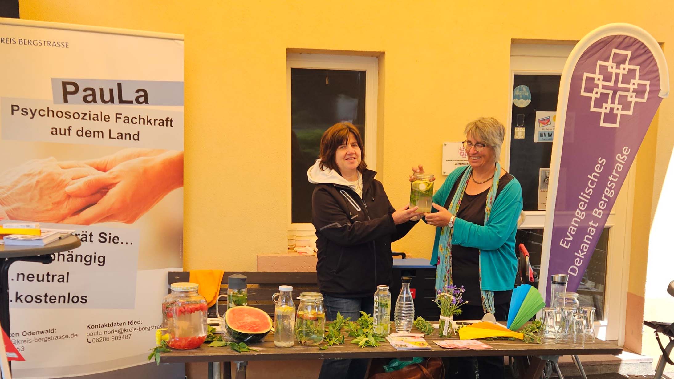Beedenkirchen_Hitzeaktionstag_C_Veranstalter_16zu9.jpg Ein Informationsstand befindet sich vor einem Gebäude. Zwei Frauen stehen hinter einem Tisch und halten gemeinsam ein Glas mit einem Getränk aus Kräutern und Früchten. Auf dem Tisch stehen mehrere Gläser und Karaffen mit aromatisiertem Wasser, frische Kräuter, eine aufgeschnittene Wassermelone sowie Flyer. Links und rechts rahmen Banner von sozialen und kirchlichen Einrichtungen den Stand ein.