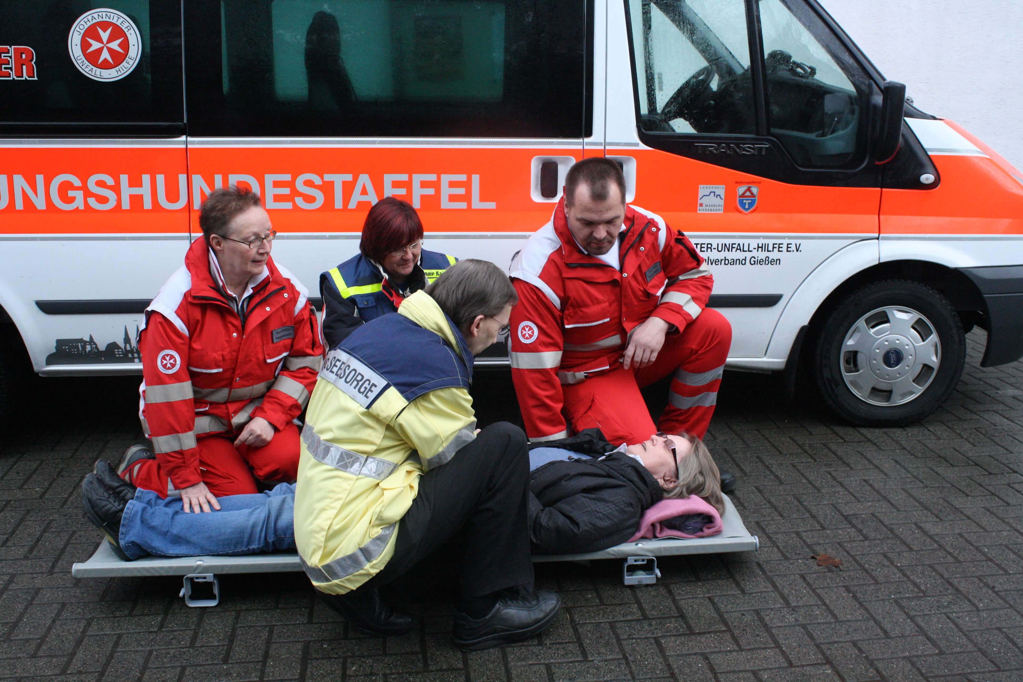 Auf dem Bild ist ein Notfallseelsorger zu sehen, der sich gemeinsam mit Rettungskräften von den Johannitern um eine auf einer Trage liegende Person kümmert.