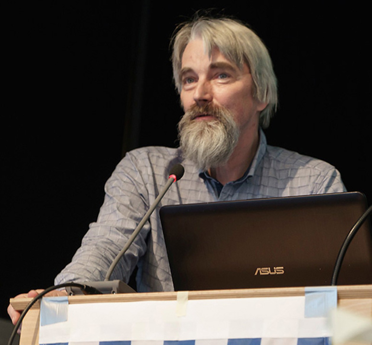 Ulrich Schneider, graue Haare, grauer Vollbart, steht an einem Rednerpult, die Hände aufgestützt, vor sich ein Notebook und ein Mikrofon. Der Hintergrund ist schwarz.