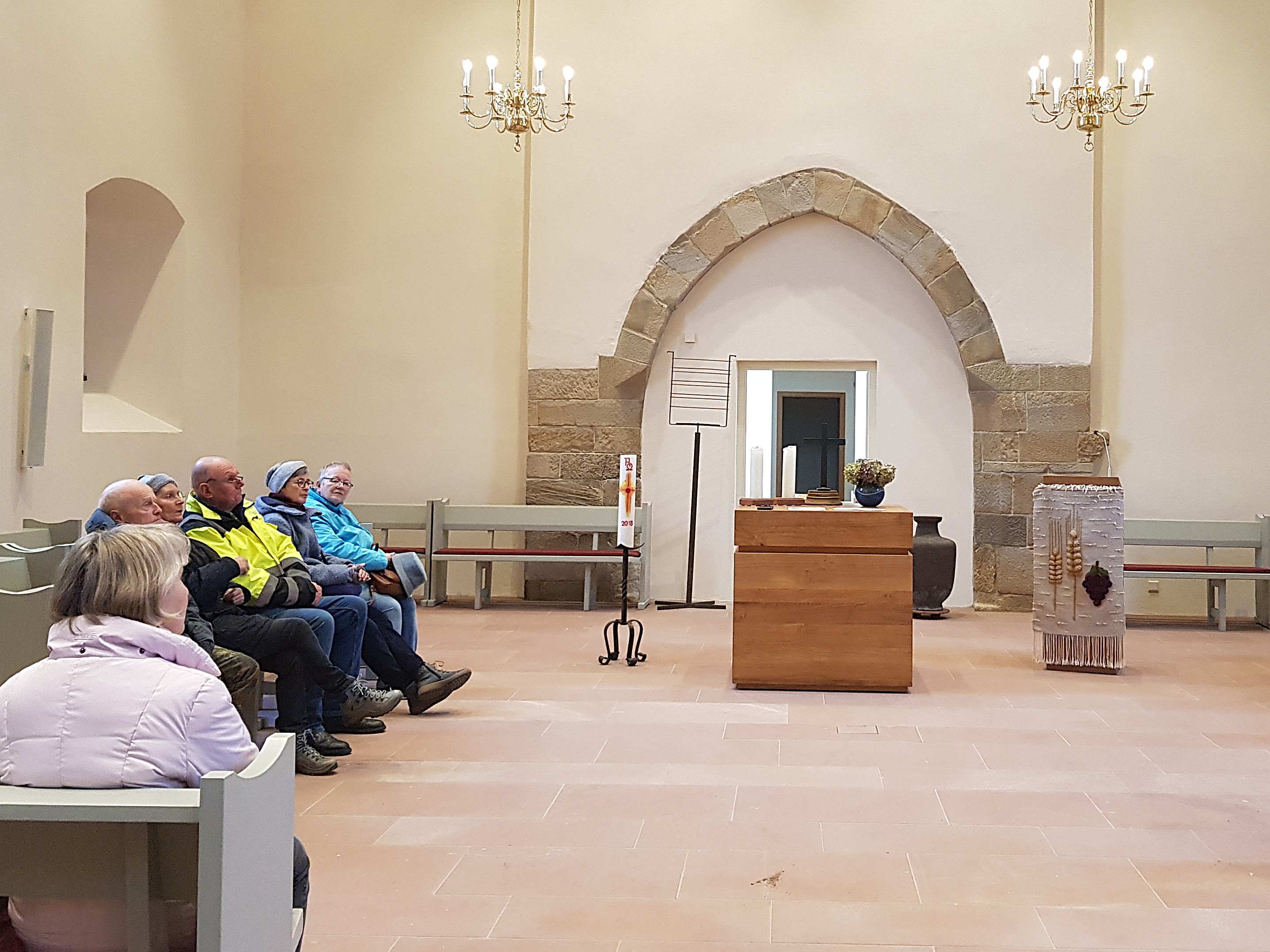 Blick in den Innenraum der Evangelischen Kirche Flomborn. Auf der linken Seite sitzen Menschen in einer Bankreihe. Mittig sieht man den Altar und Kerzen.