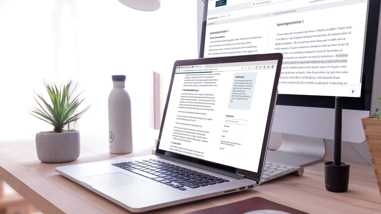 Heller, aufgeräumter Arbeitsplatz mit einem geöffneten Laptop vor einem großen Monitor. Auf beiden Bildschirmen ist Text dargestellt. Neben dem Laptop stehen eine Wasserflasche und eine kleine Pflanze in einem Topf auf einem Holztisch.