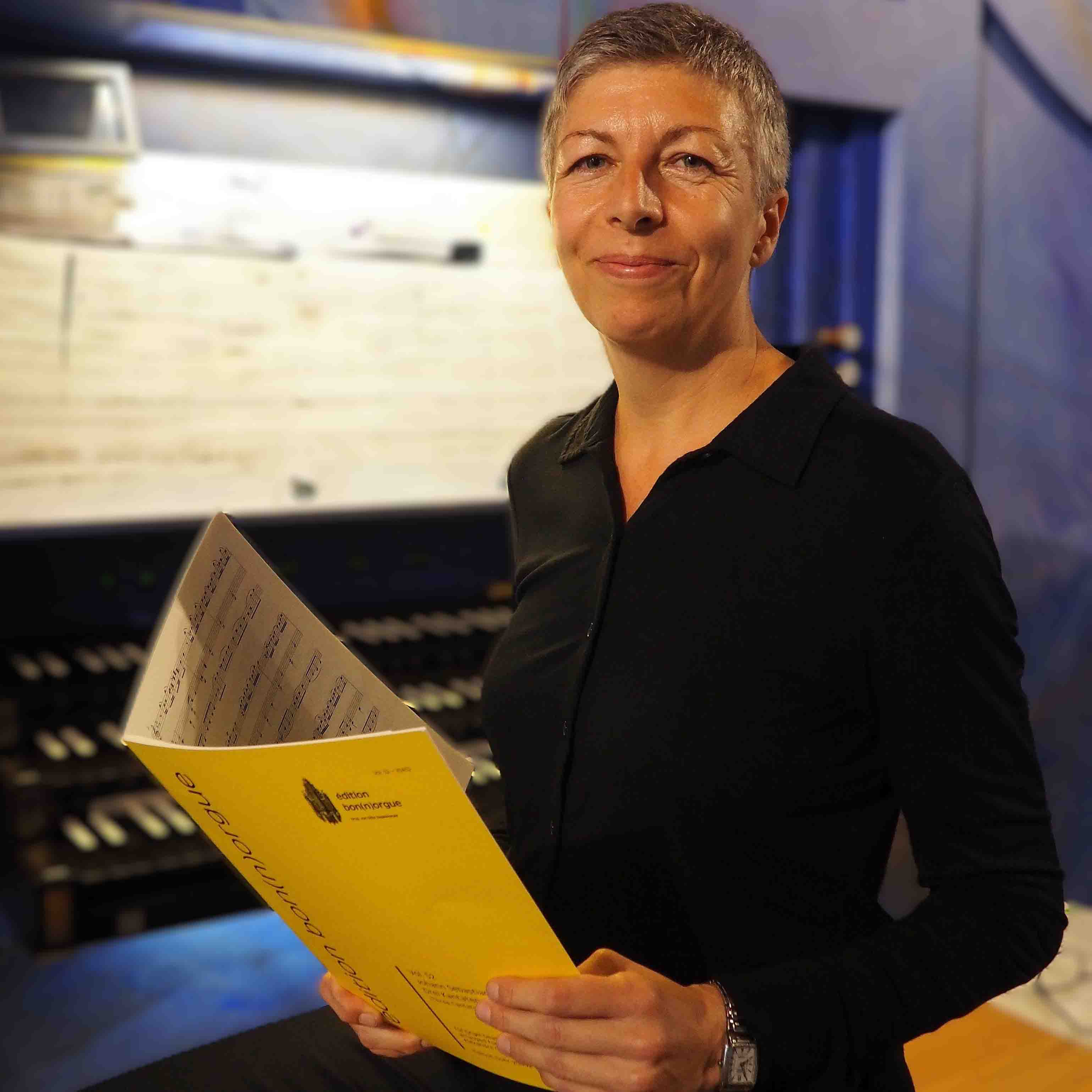 Lächelnde Frau mit kurzen grauen Haaren hält gelbe Notenblätter vor einer Orgel.