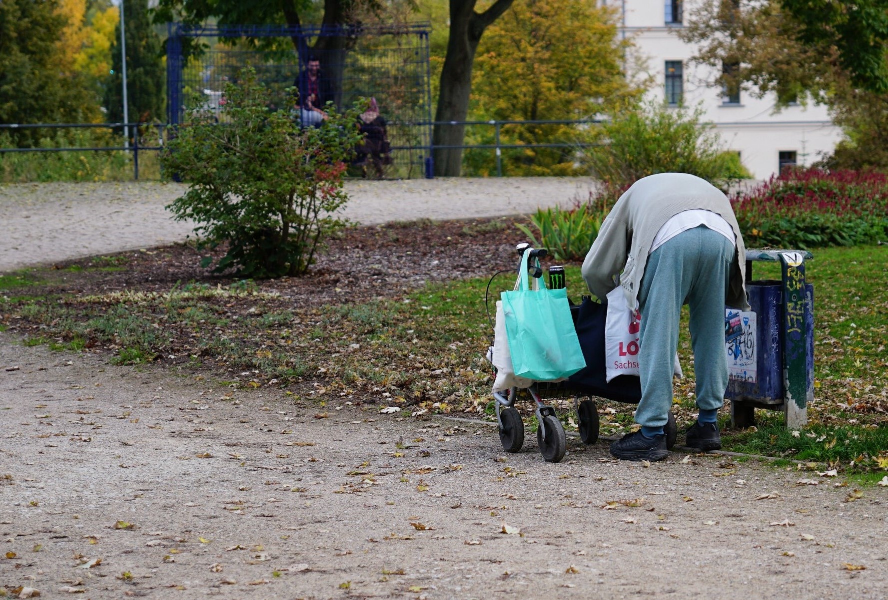 Ein menschenleerer Park im Herbst. Ein Mann in ärmlicher Kleidung steht gebückt mit dem Rücken zum Betrachter , vor sich einen Rollator, an dem viele Taschen und Beutel hängen. Offensichtlich sucht er in einem Abfallbehälter neben einer Bank nach etwas Verwertbarem.