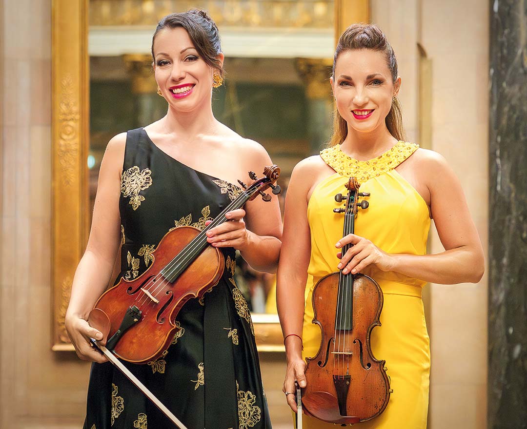 Zwei lächelnde Musikerinnen in eleganter Abendkleidung halten jeweils eine Violine und stehen nebeneinander in einem festlichen Innenraum.