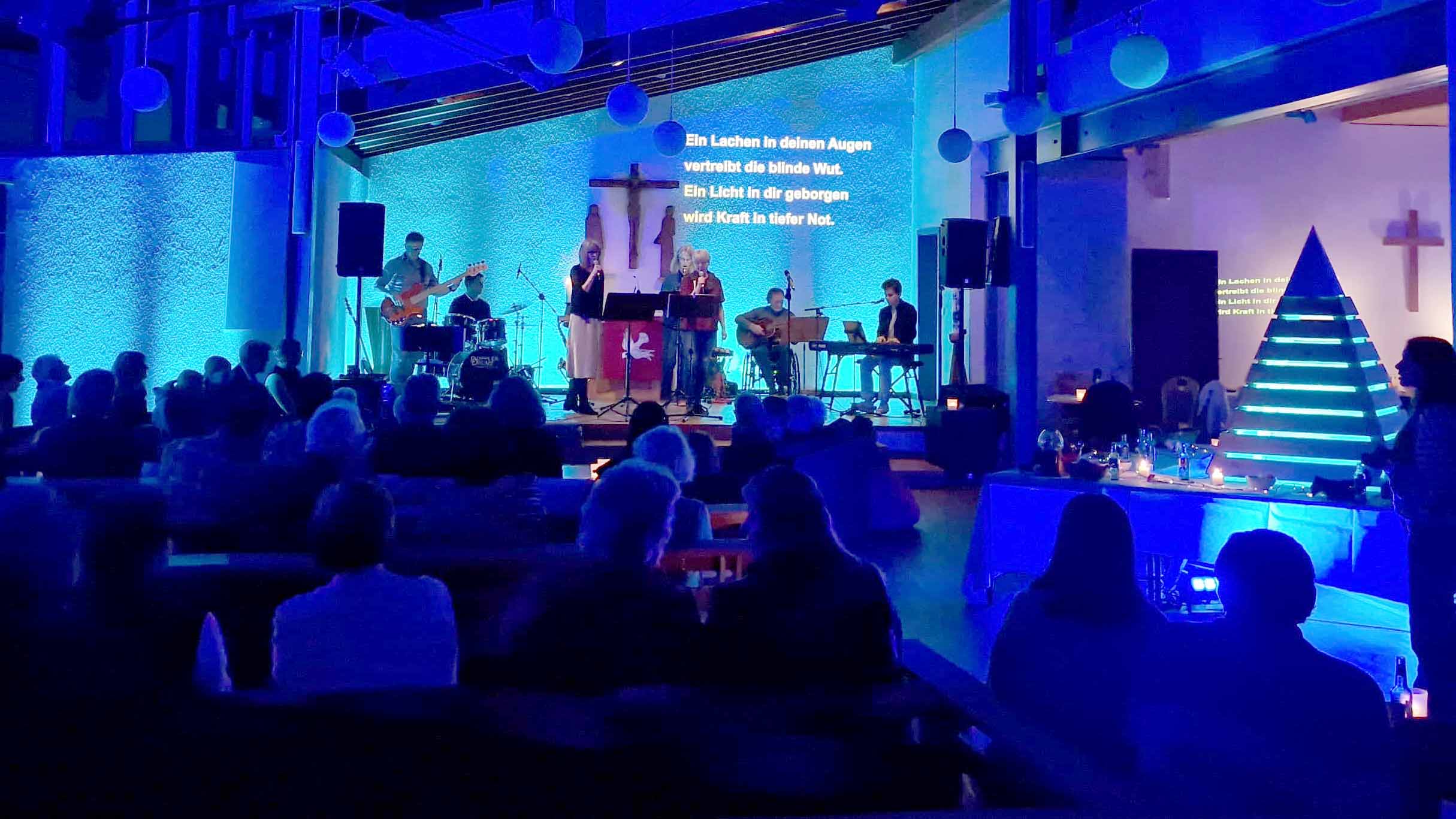 Bensheim_Stephanus_Wohnzimmerkirche_C_Veranstalter_16zu9.jpg Blick in die blau und türkis beleuchtete Stephanuskirche in Bensheim während einer Wohnzimmerkirche. Auf der Bühne musiziert eine Band mit Gesang, Gitarren, Schlagzeug und Keyboard. Zahlreiche Besucherinnen und Besucher verfolgen die Veranstaltung von den Kirchenbänken aus. An der Wand hinter der Band sind Liedtexte eingeblendet. Rechts steht eine pyramidenförmige Lichtinstallation, davor laden gedeckte Tische mit Kerzen zum Verweilen ein.
