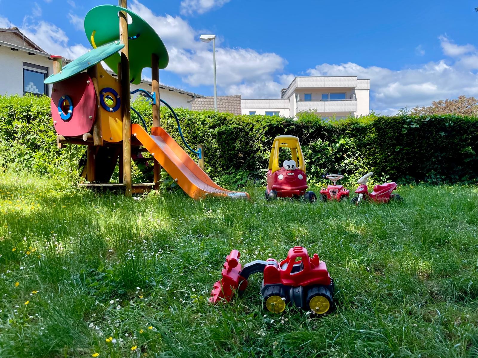 Das Bild zeigt einen kleinen Spielplatz in einem Garten. Im Vordergrund befindet sich ein roter Spielzeugbagger im Gras. Im Hintergrund sind eine bunte Rutsche und mehrere kleine Spielzeugautos zu sehen. Der Garten ist grün und gepflegt, mit einer Hecke, die ihn umgibt. Im Hintergrund sind Wohnhäuser unter einem blauen Himmel mit einigen Wolken sichtbar.
