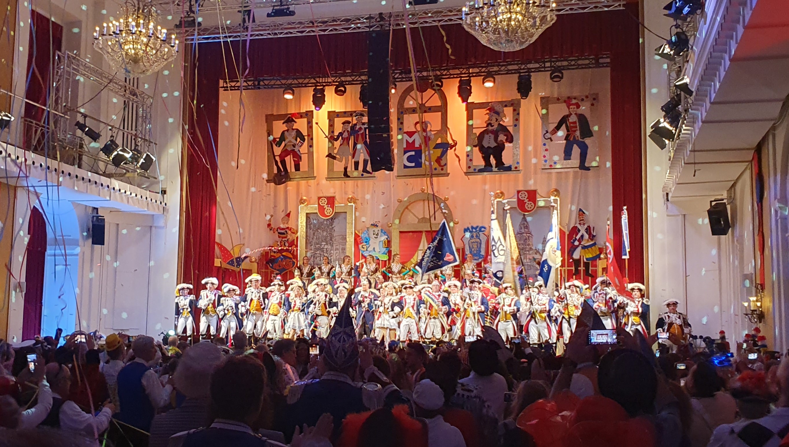 Blick auf die geschmückte Bühne, vor dem Elferrat stehen viele Mitglieder der Ranzengarde. Davor Publikum.