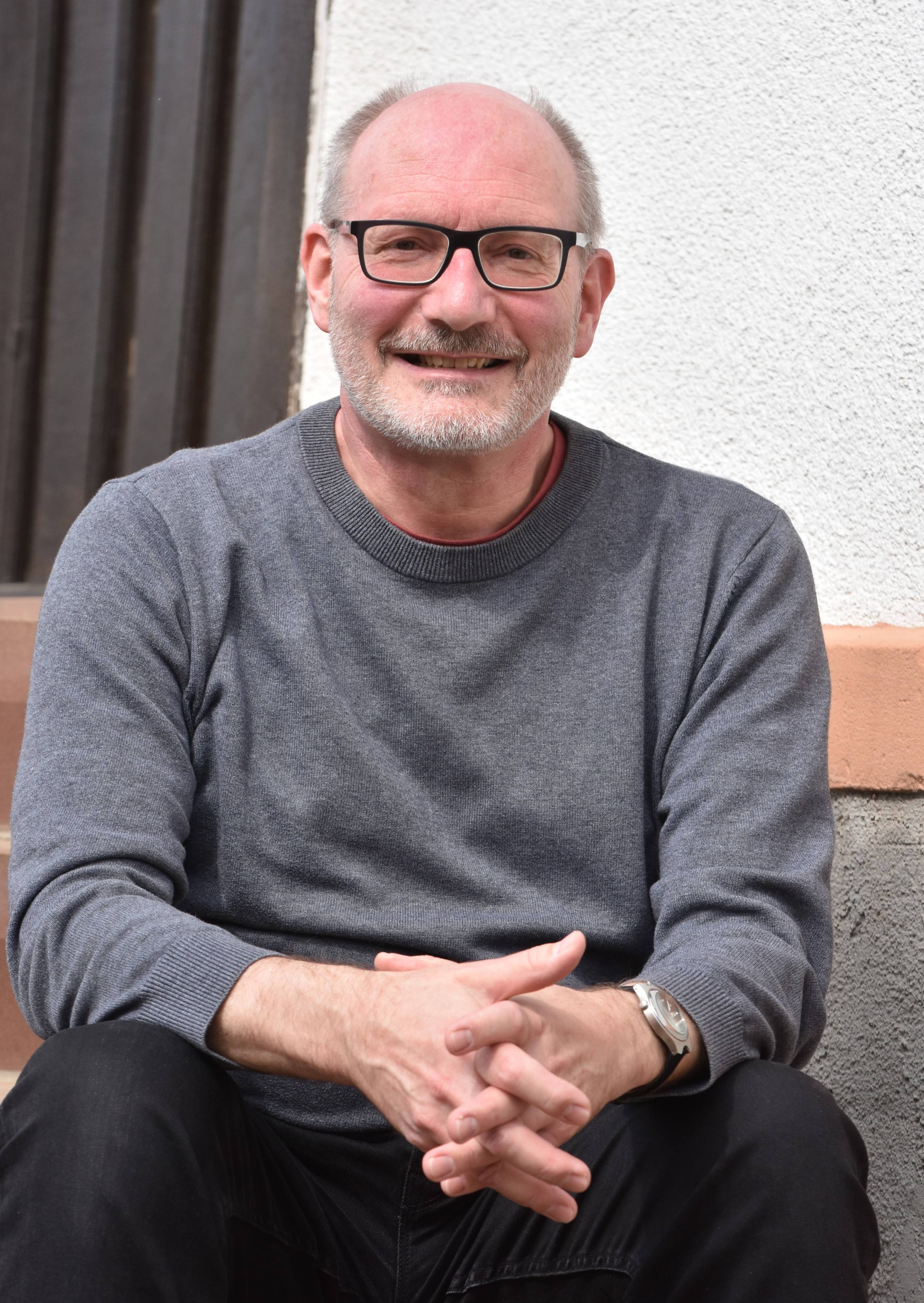Burkhard Zentgraf, Brille, Bart, grauer Pullover und schwarze Jeans, sitzt auf einer Treppe, hat die Arme auf den Oberschenkeln liegen und faltet die Hände, schaut zufrieden aus.