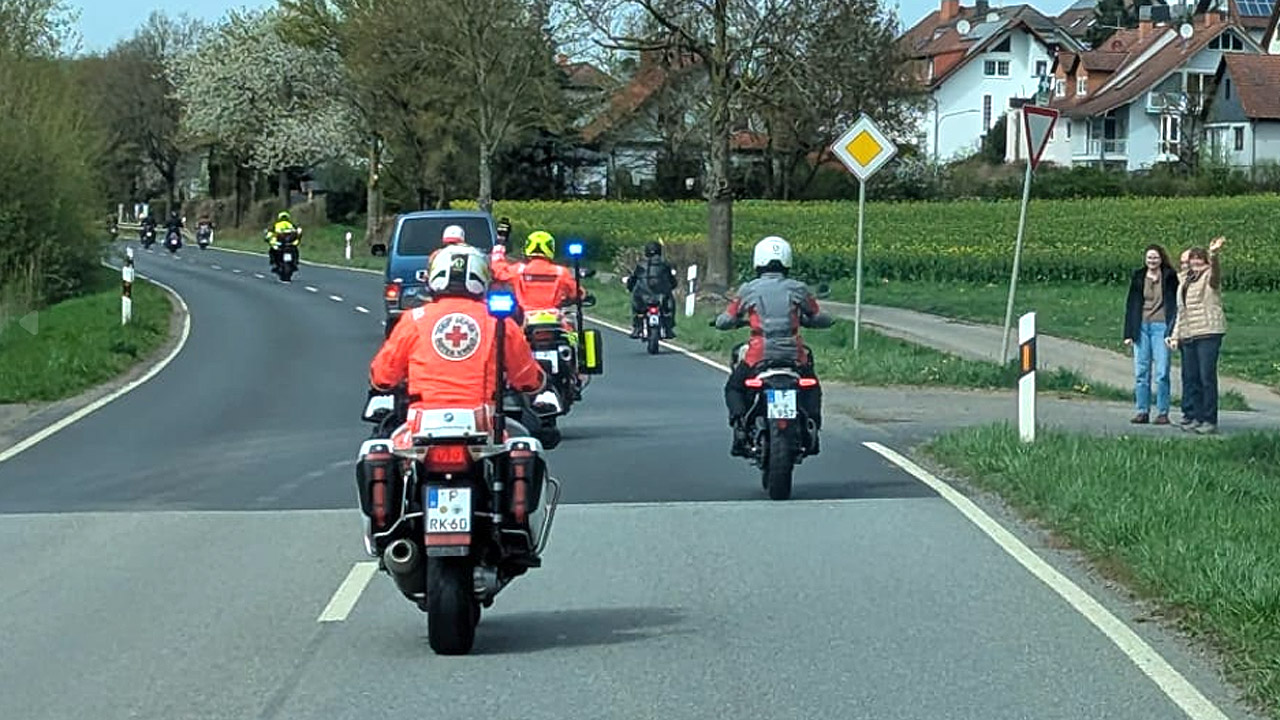 Gruppe von Motoarradfahrern fährt in einen Ort, Menschen winken am Straßenrand