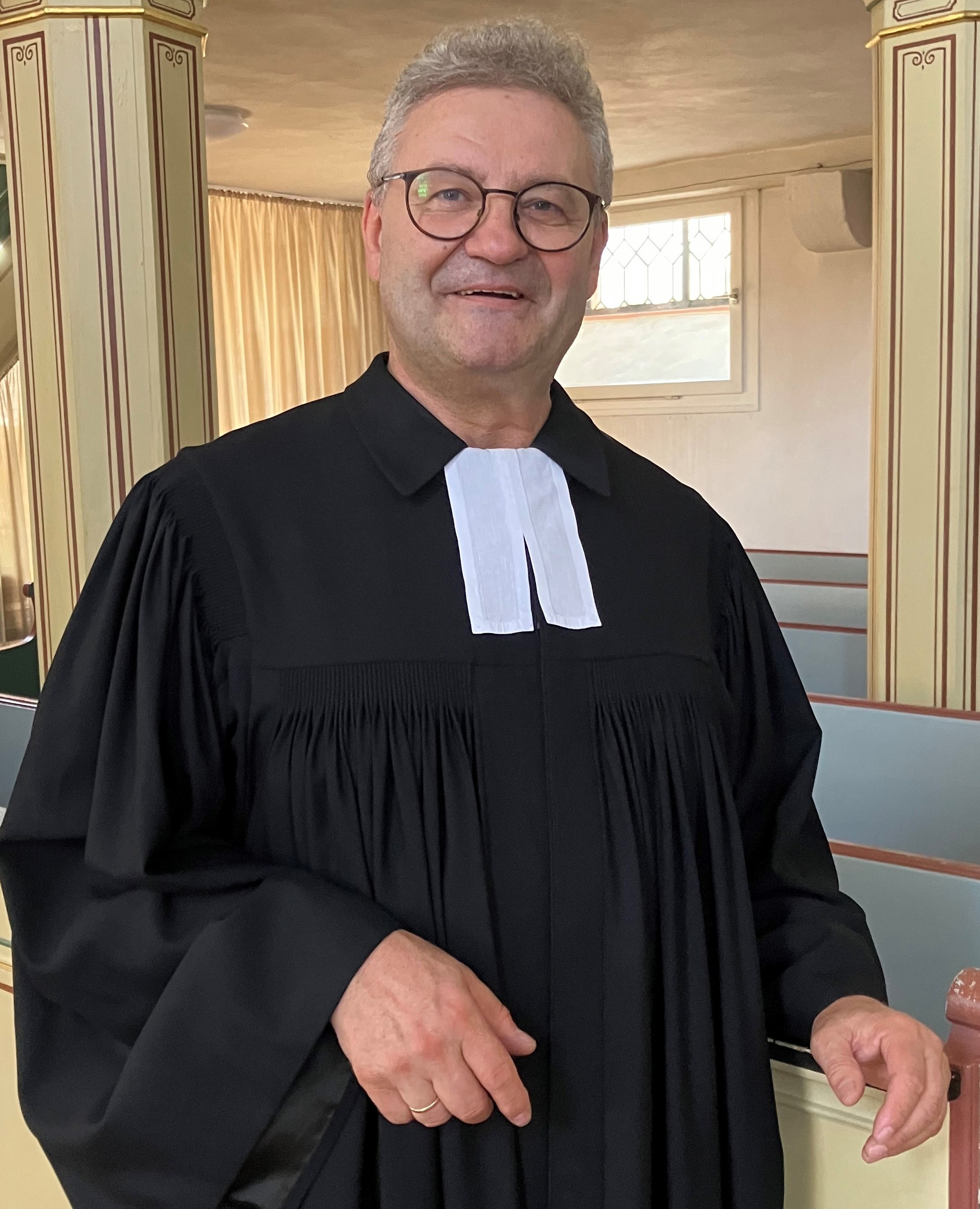 Markus Christ, im Talar, kurze graue Haare, Brille, steht in Langen-Bergheim im Kirchenraum, die Hände hält er locker vor dem Körper.