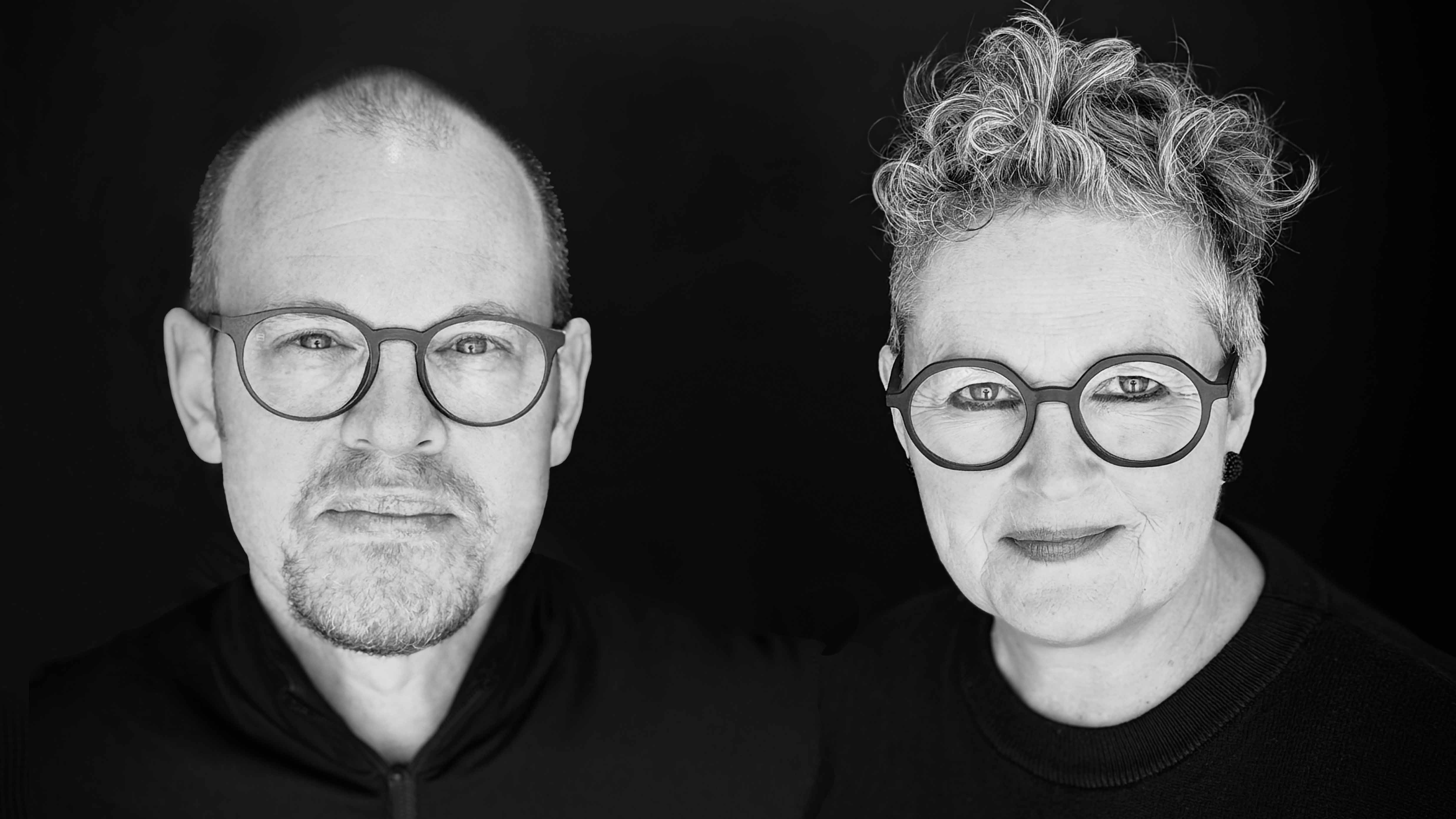 Schwarz-Weiß-Porträt eines Musikduos vor schwarzem Hintergrund. Links ein Mann mit kurzem Haar, rundlicher Brille und Kinnbart, rechts eine Frau mit lockigem, hochgestecktem Haar und großer runder Brille. Beide tragen dunkle Oberteile und blicken direkt in die Kamera.