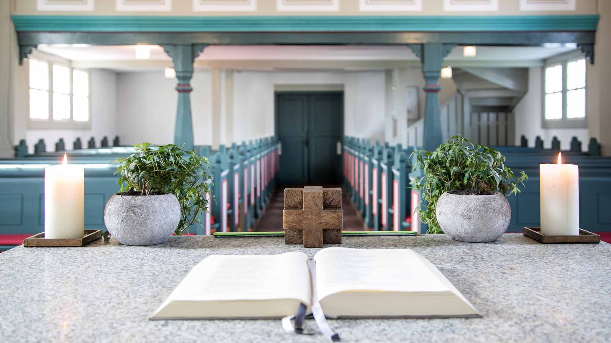 Offenes Buch und Holzkreuz auf dem Altar einer hellen Kirche, eingerahmt von zwei Kerzen und zwei Topfpflanzen; dahinter leere Kirchenbänke.