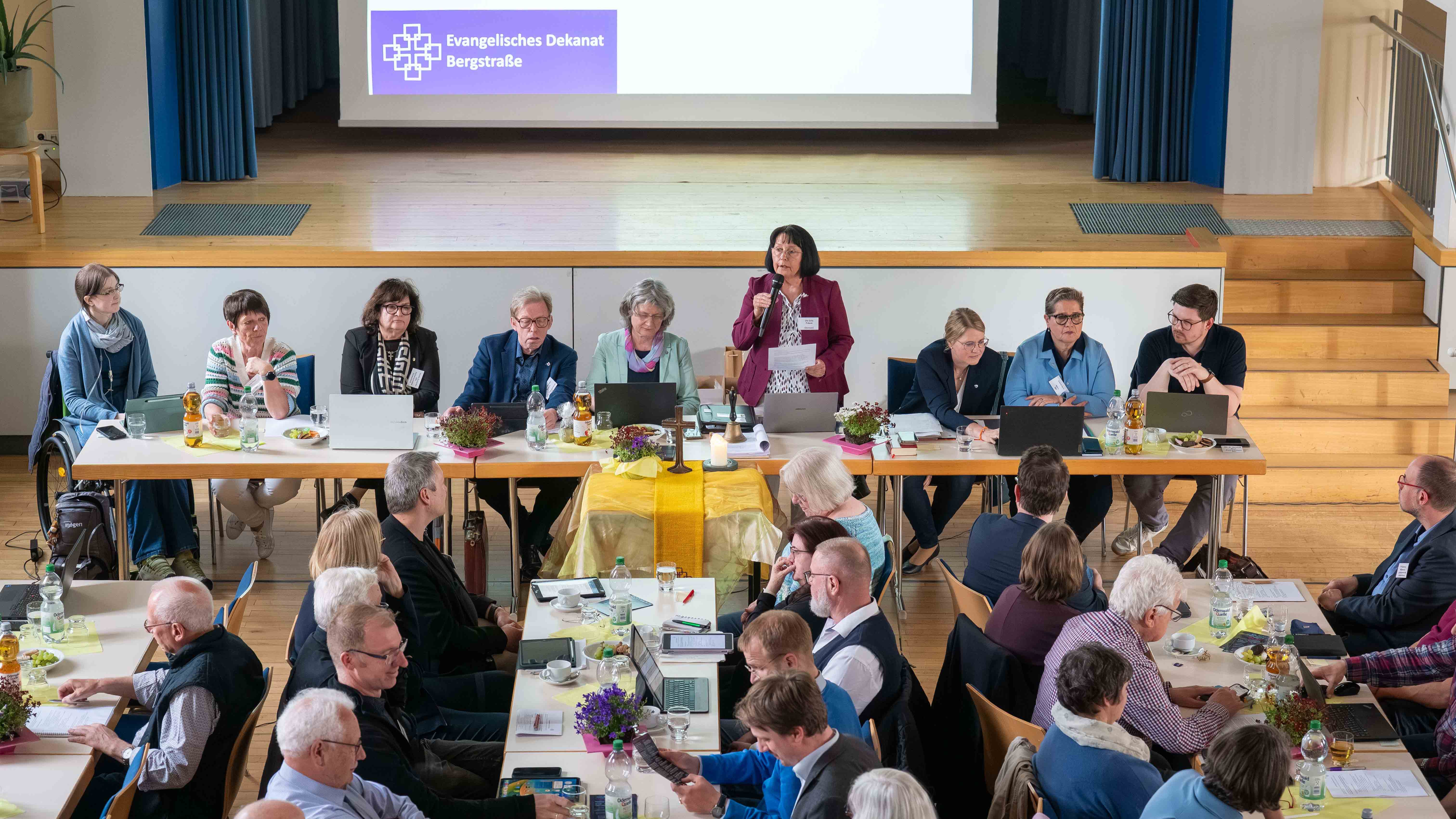 Eine Frau - Präses Ute Gölz - spricht bei der Synode des Evangelischen Dekanats Bergstraße am Vorstandstisch ins Mikrofon. Vor ihr sitzen die Synodalen an langen Tischreihen.