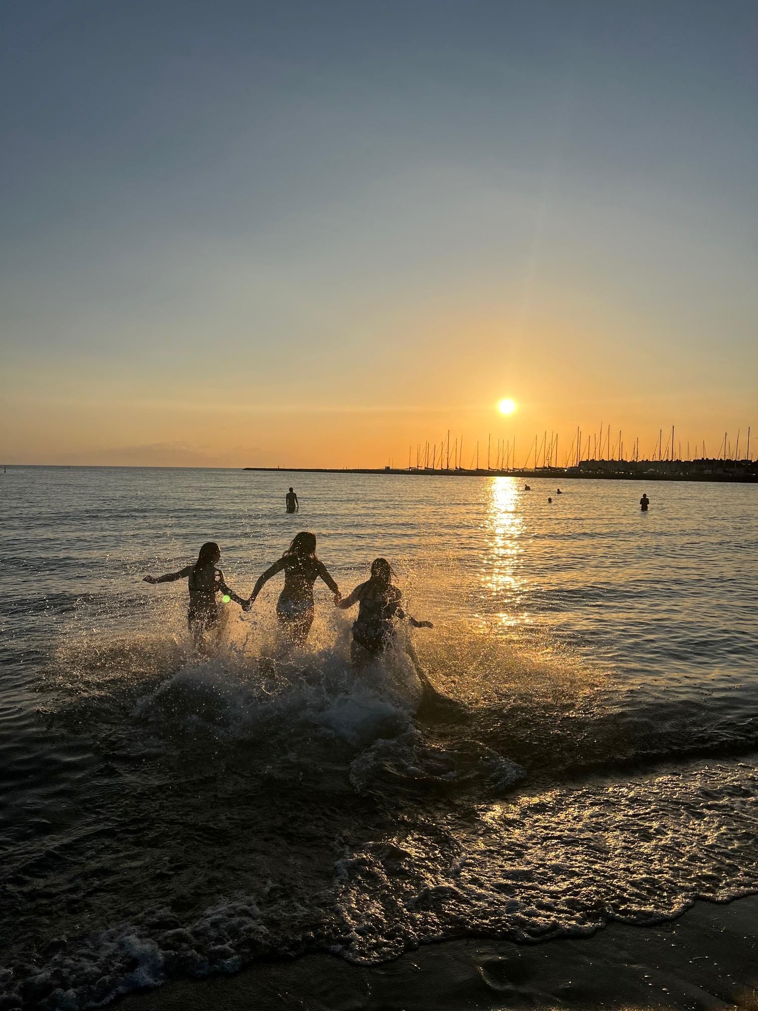 Sonnenuntergang am Meer, die Sonne steht leuchtend kurz über dem Horizont und taucht ihn in orange-gelbes Licht, man sieht drei junge Menschen von hinten, die sich an den Händen halten und durchs Wasser laufen, davor Wellen