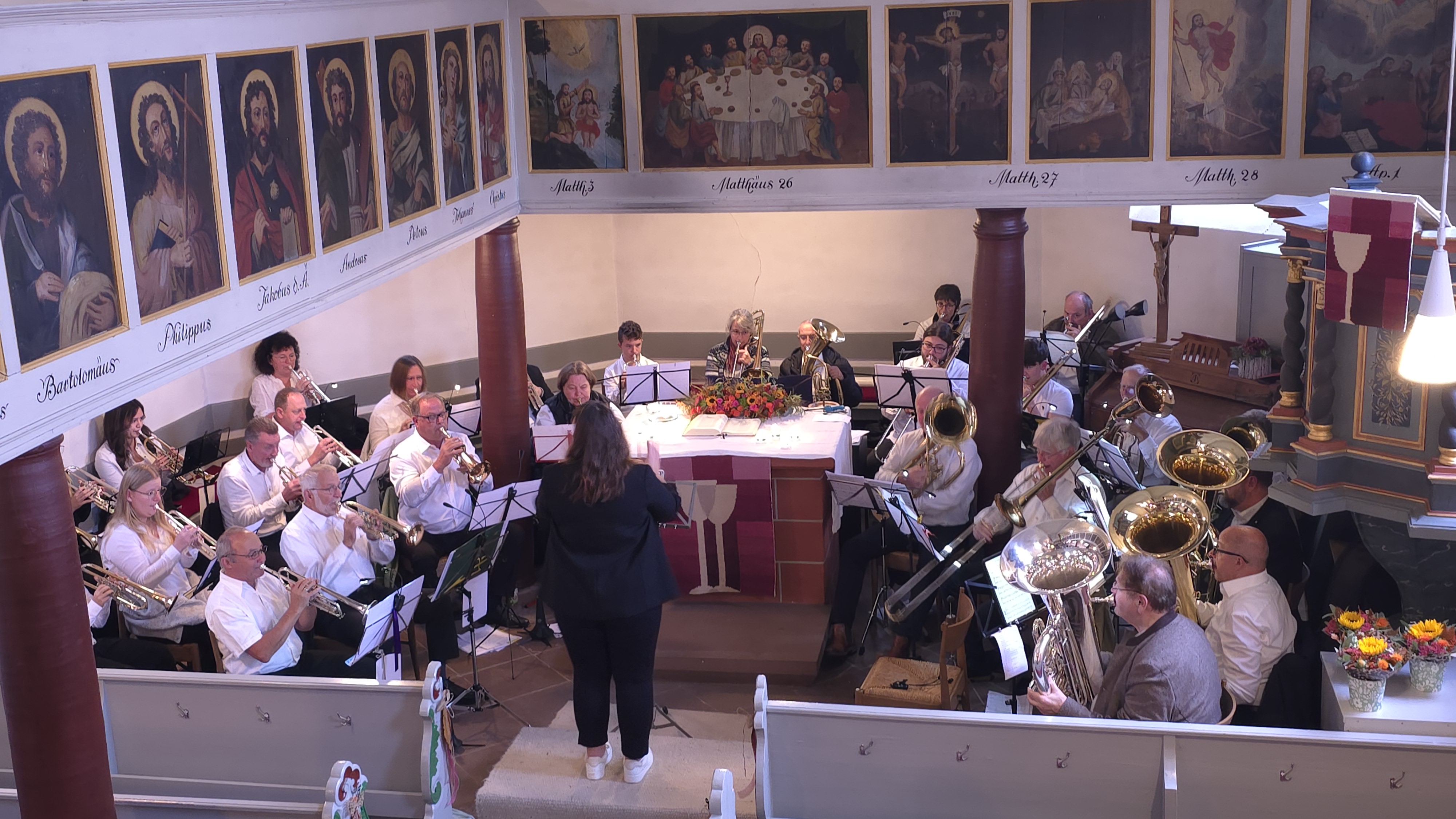 Blick in den Alterraum der Kirche Wallernhausen, dicht gedrängt sitzt eine Gruppe von etwa 30 Blechbläsern rund um den Altar, dirigiert von einer jungen Frau.