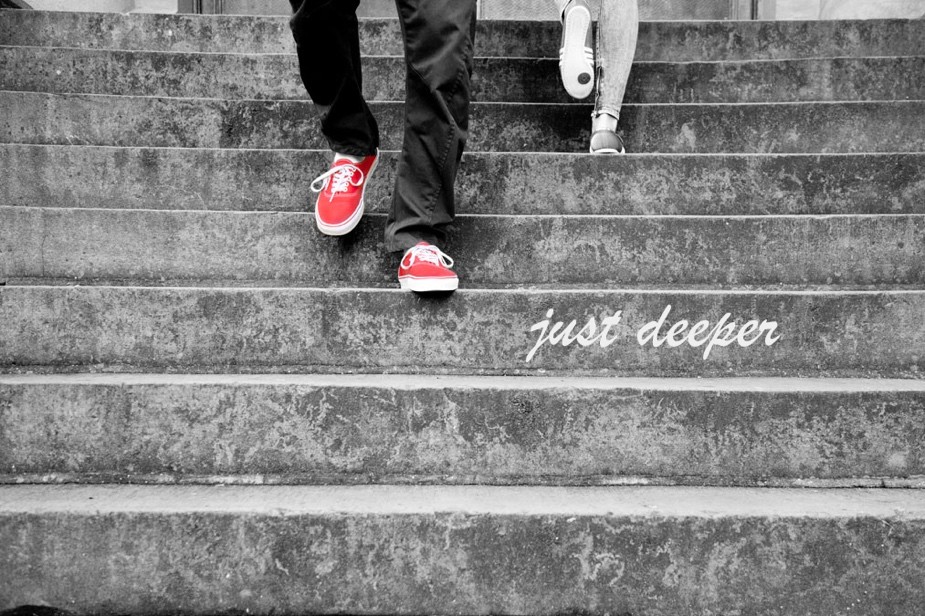 Das Bild zeigt eine Schwarz-Weiß-Aufnahme von zwei Personen, die eine Treppe hinuntergehen. Die vordere Person trägt dunkle Jeans und rote Turnschuhe, die ein Farbtupfer in dem ansonsten monochromen Bild sind. Die Person hinter ihr scheint helle Schuhe zu tragen. Auf einer der Stufen ist der Text „just Deeper" geschrieben.