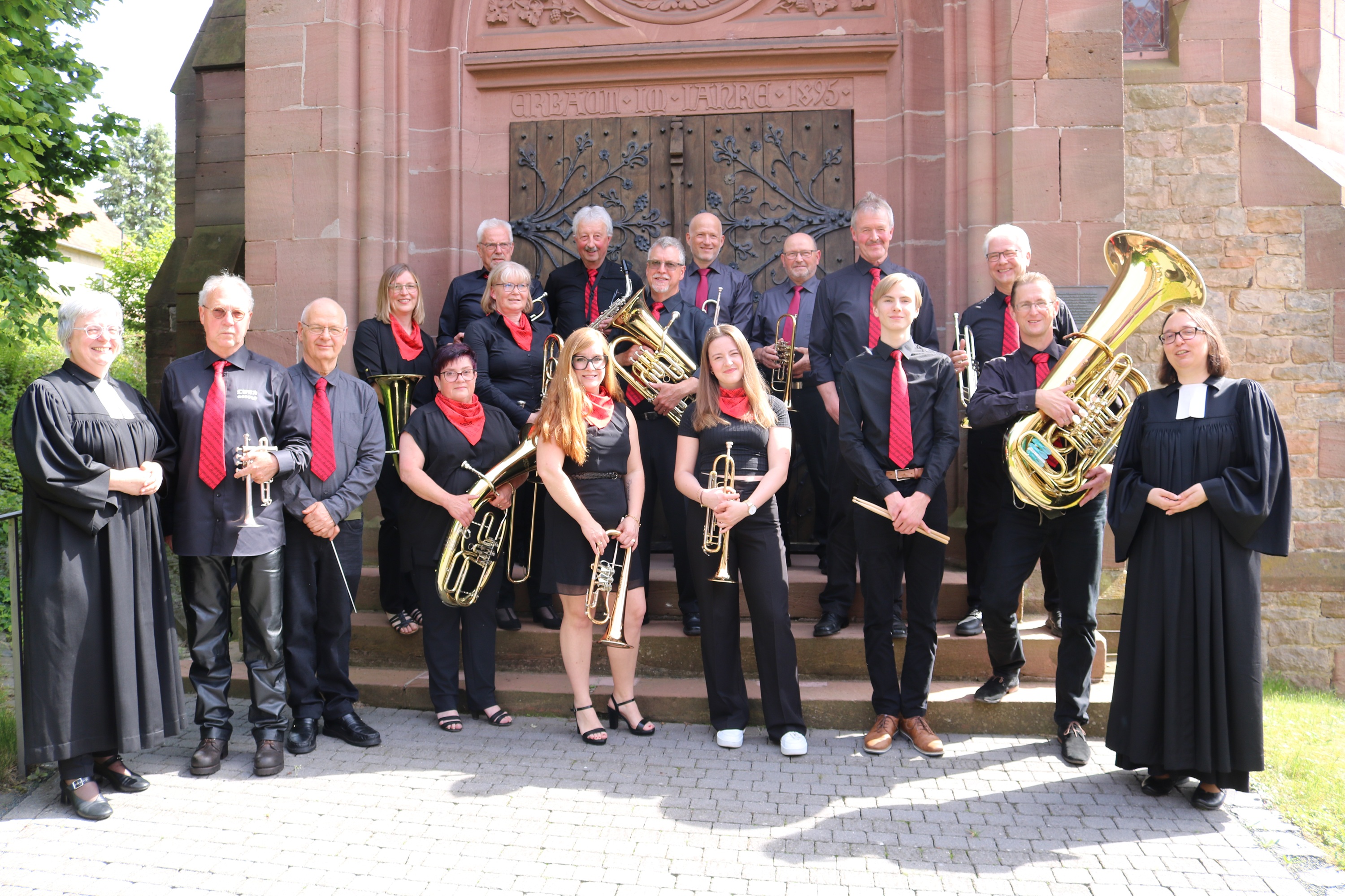 Menschen mit Blasinstrumenten vor einer Kirche, rechts und links eine Pfarrerin