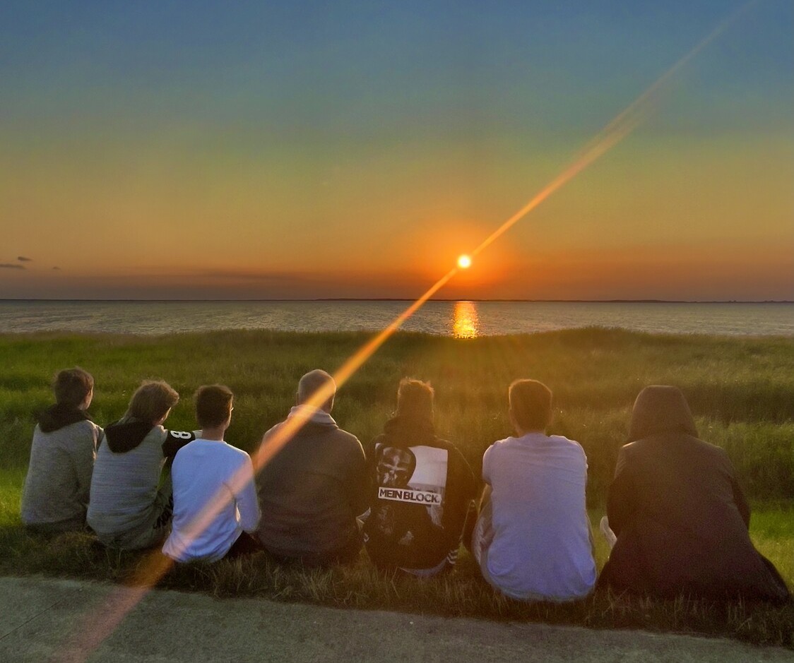 Das foto zeigt eine Gruppe Jugendlicher (sieben Personen), die mit dem Rücken zur Kamera am Boden einer Strandpromenade sitzen, übers Meer schauen und den Sonnenuntergang beobachten. Die Sonne steht in leuchtendem Orange kurz über dem Horizont.