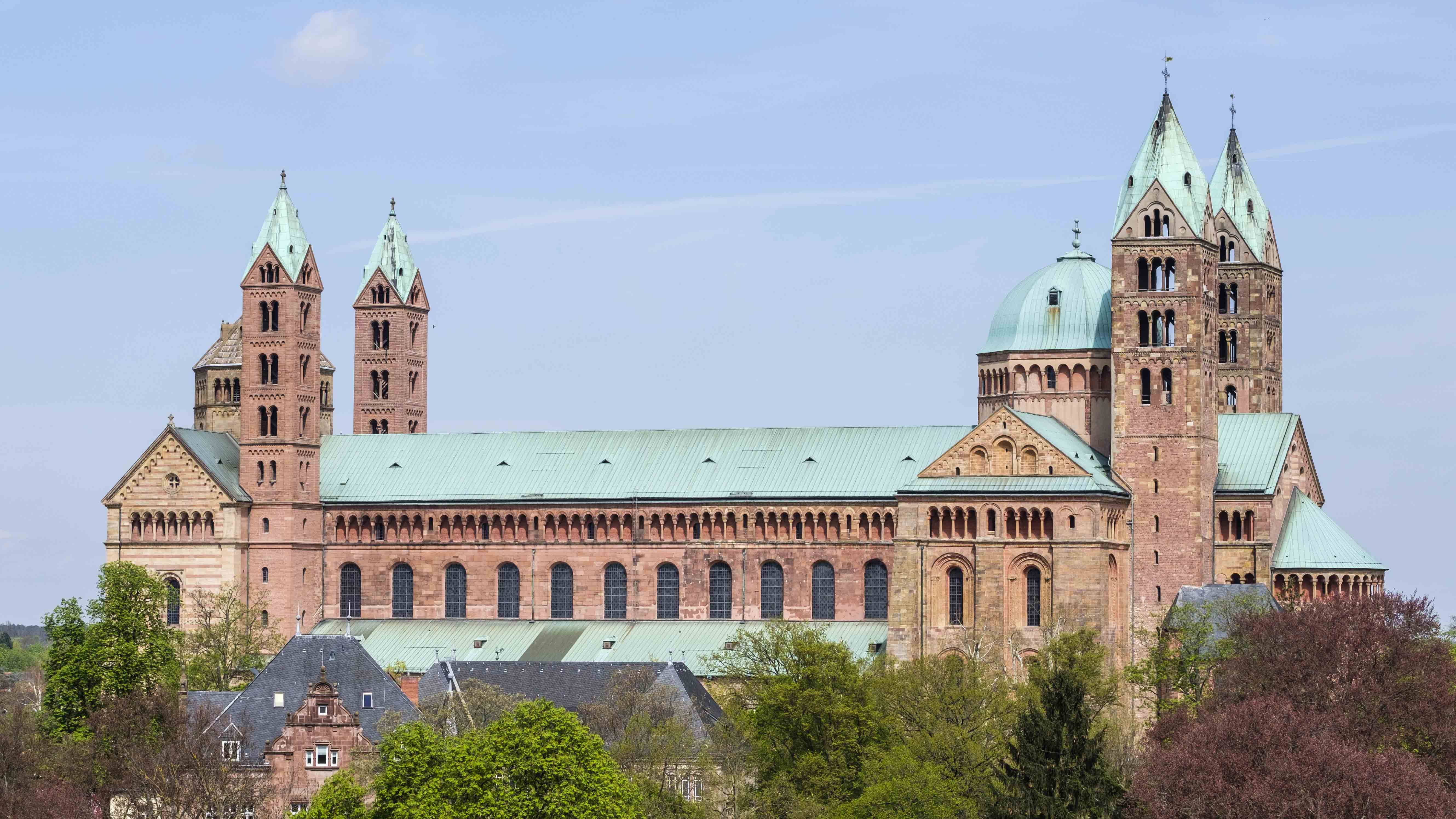 Romanischer Dom aus rotem Sandstein mit vier Türmen und grünen Kupferdächern, umgeben von Bäumen und kleineren Gebäuden.