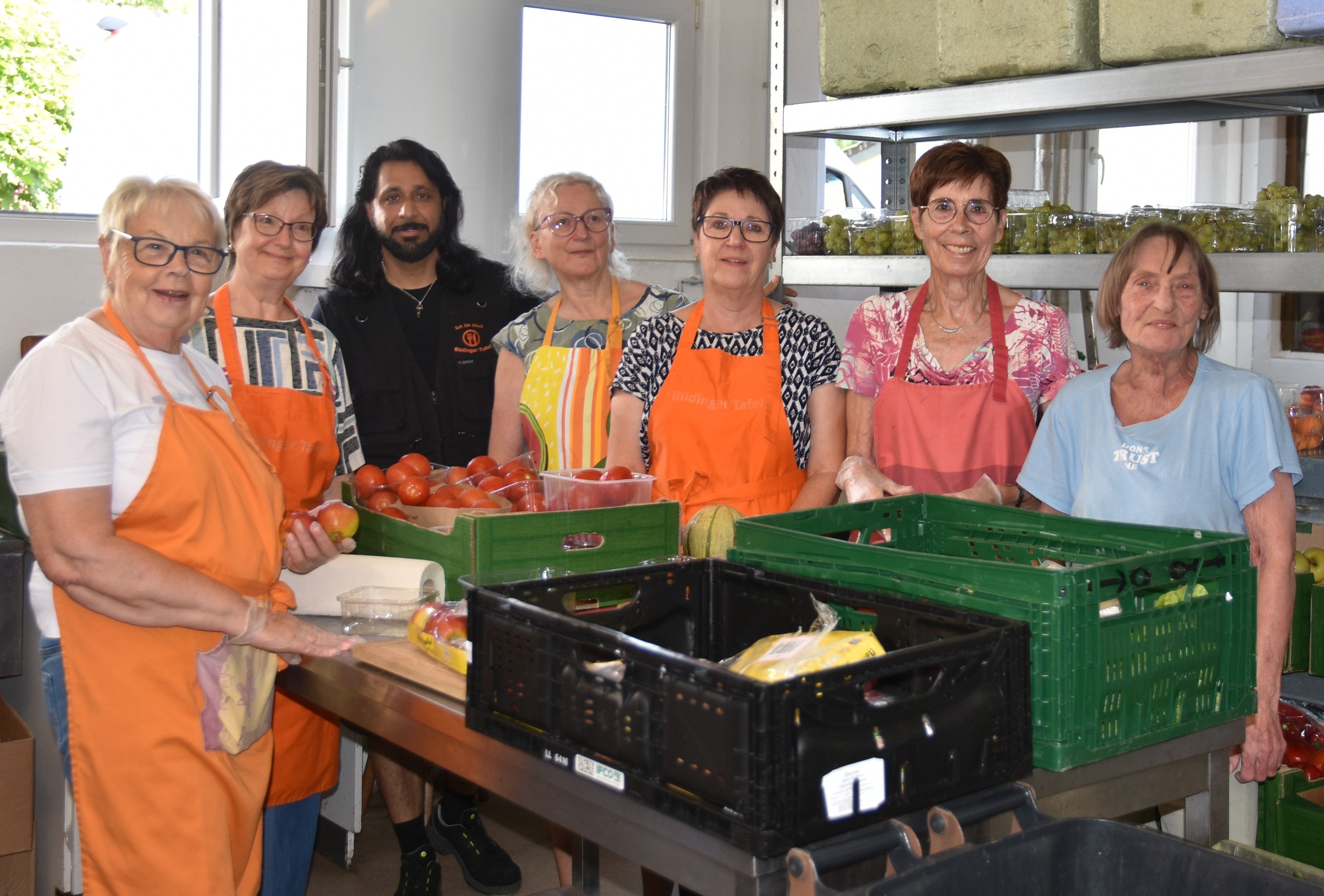 Sechs Frauen und ein Mann, viele tragen eine orangefarbene Schürze, stehen um einen Tisch, auf dem Kisten mit Lebensmittel stehen, dahinter Regale, ebenfalls mit hauptsächlich Obst gefüllt. Es sieht nach Arbeit aus.